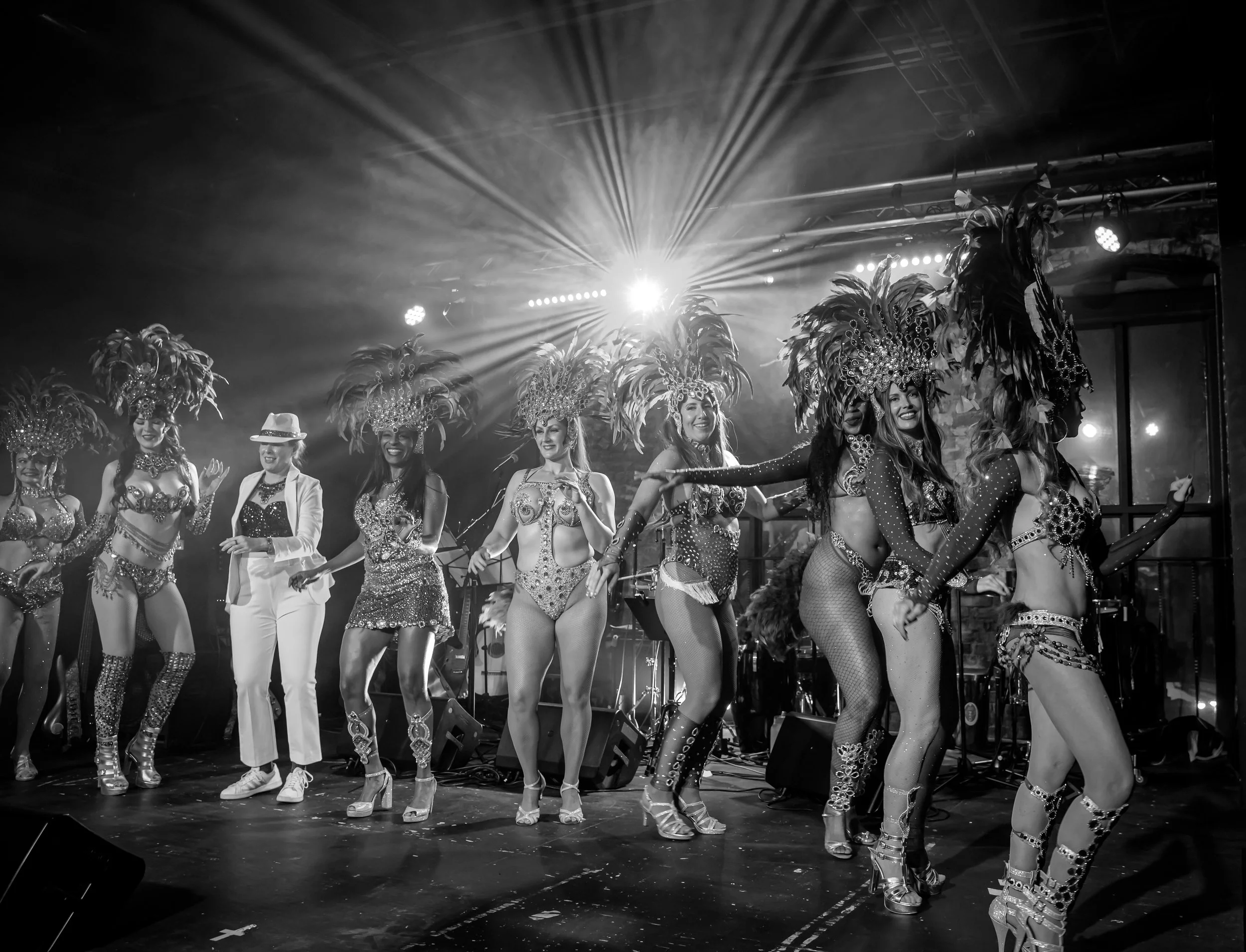 Black and white photo of showgirls on stage at Carnaval Austin wearing elaborate feathered headdresses and costumes, with stage lights in the background.