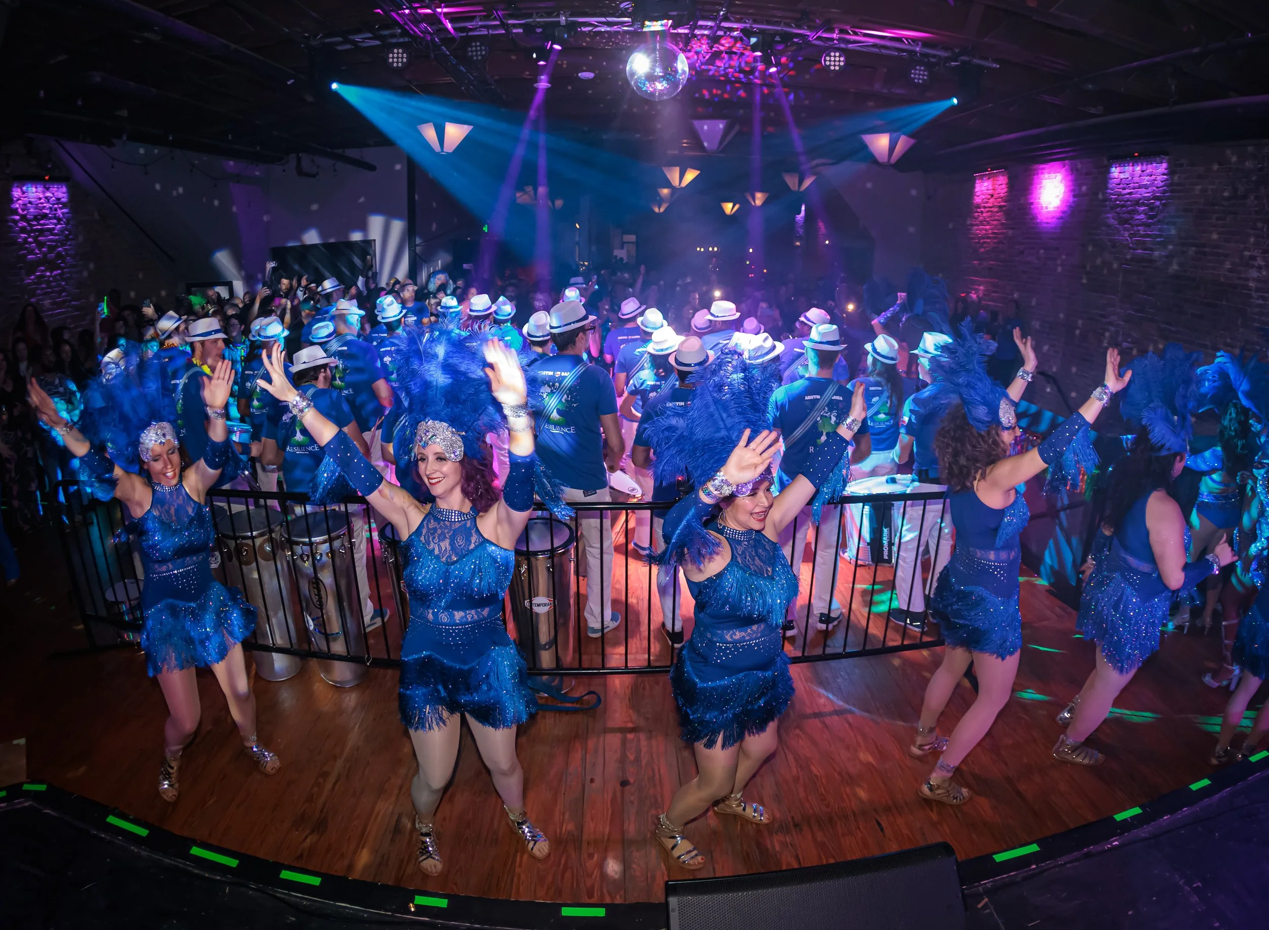 Carnaval's lively party scene with women dancing in front, wearing matching blue costumes and feathered headpieces. Behind them, a large crowd of people, many wearing hats, are dancing and enjoying the event under colorful lighting and a disco ball.