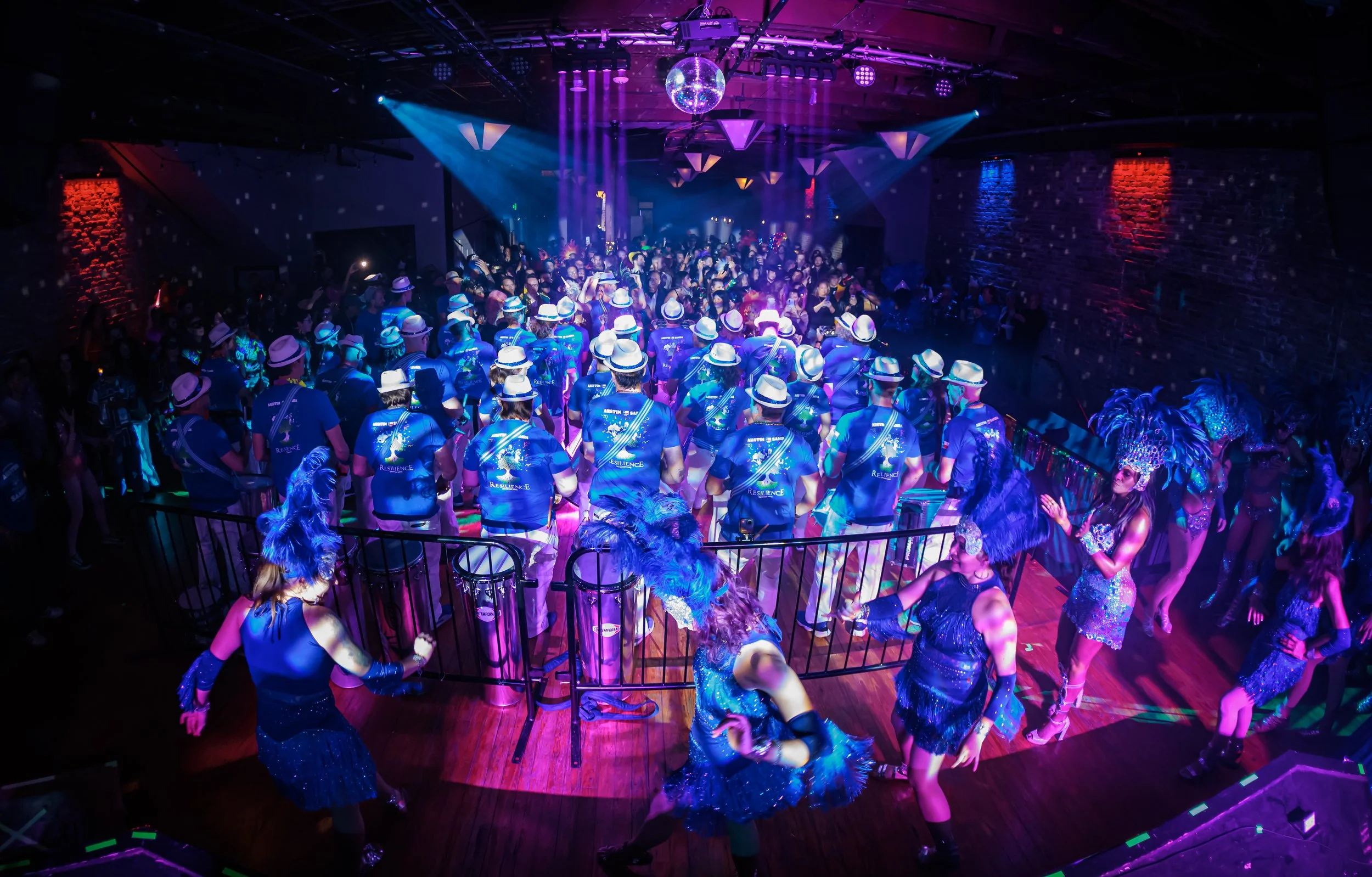 Carnaval Austin's lively dance party with a large crowd, colorful lighting, and performers in costumes with feathered headdresses on a dance floor.