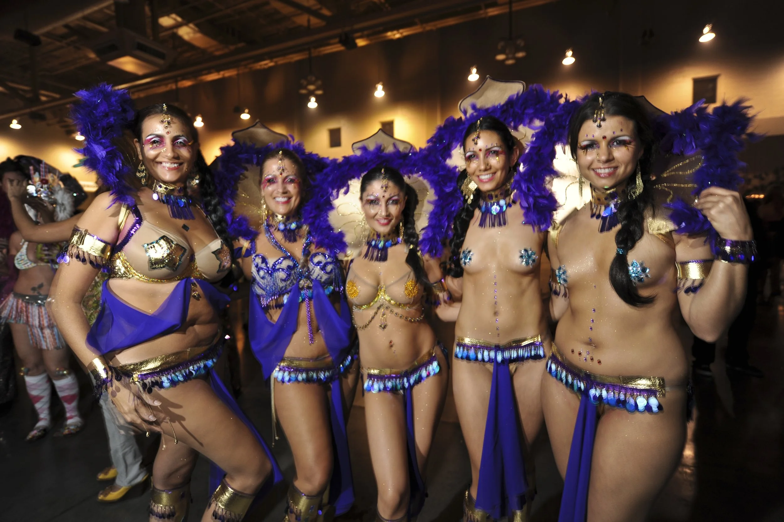 Group of five women dressed in colorful carnival costumes with feathers, sequins, and jewelry, posing together at Carnaval Austin. Returning February 7, 2026.