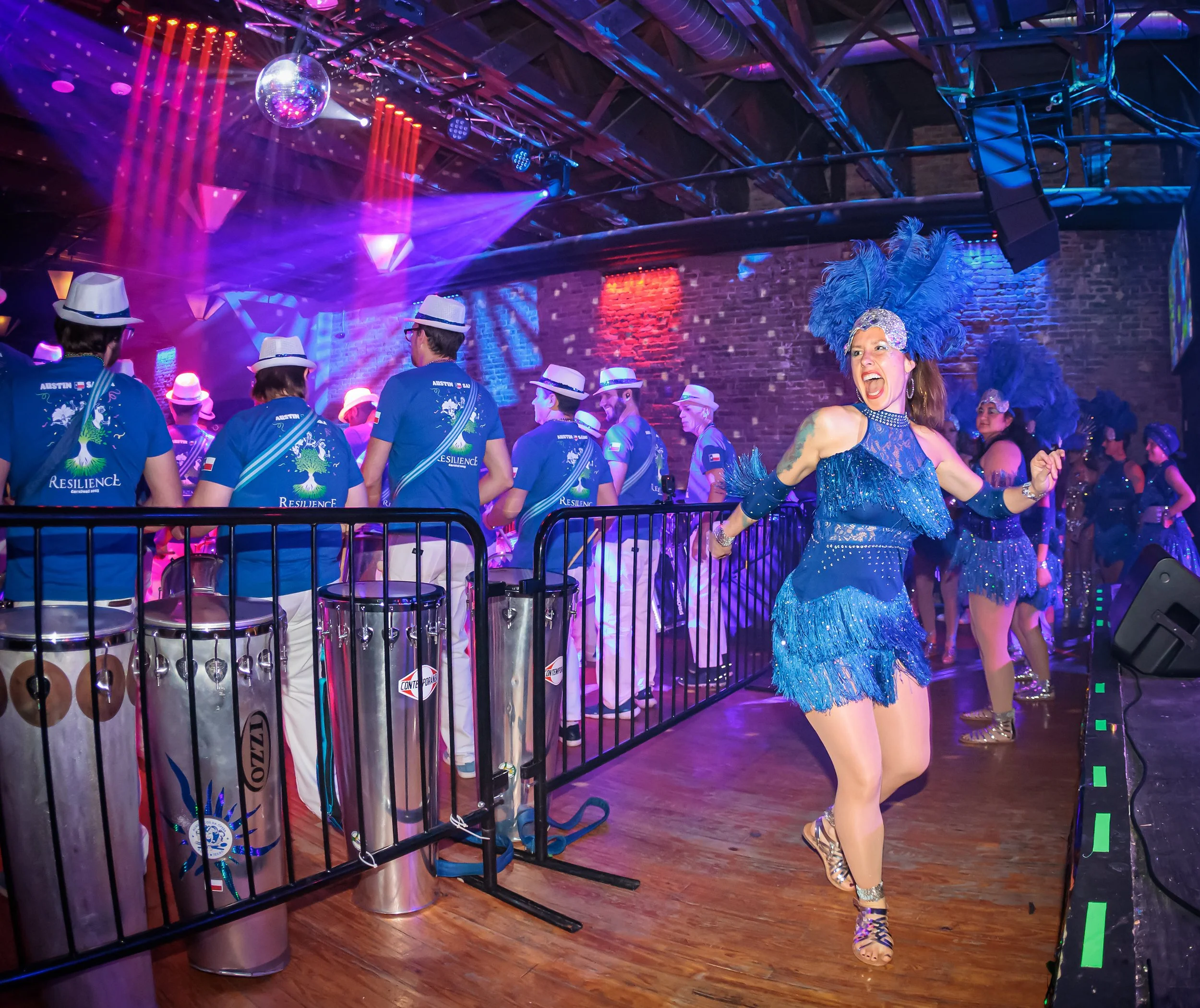 A woman dancing in a blue flapper-style outfit with feathers, in front of a band of musicians wearing blue shirts and white hats at Carnaval Austin with colorful lights and a disco ball.