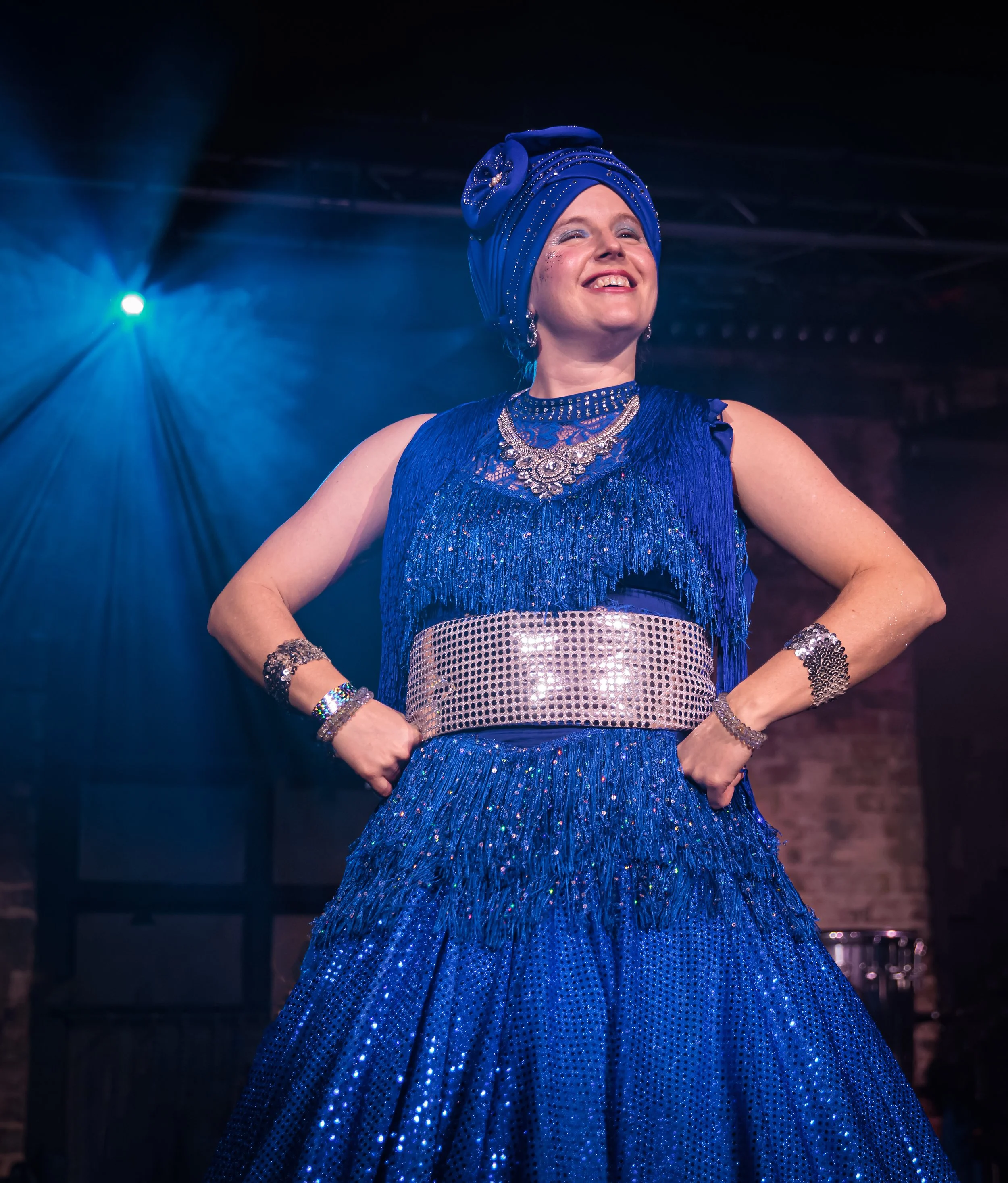 Woman smiling, wearing a vibrant blue dress with fringe and sequins, accessorized with jewelry and a matching headwrap, standing confidently on stage at Carnaval Austin with blue lighting. Returning February 7, 2026.