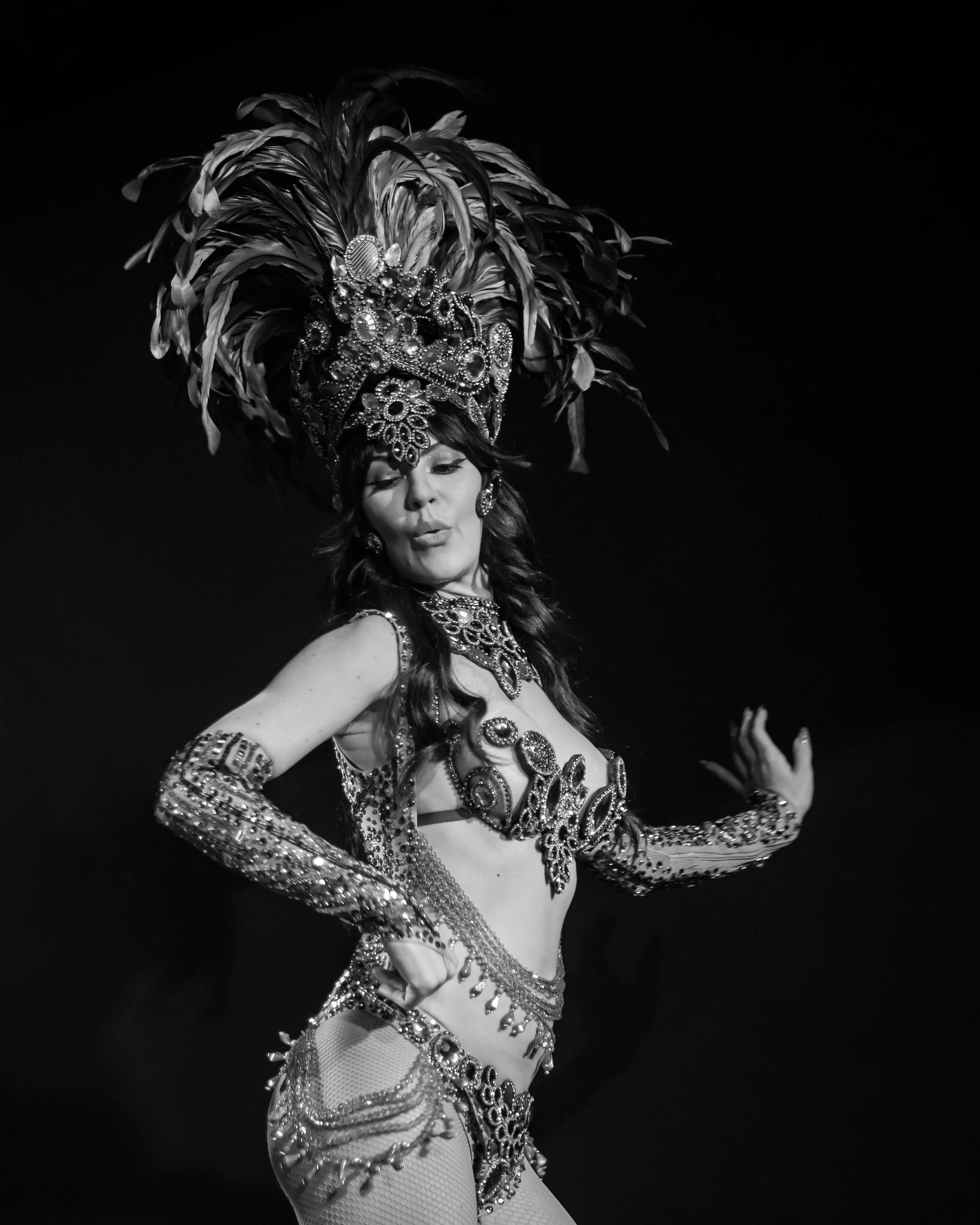 A woman dressed in a showgirl costume with a large feathered headdress and jewel embellishments, striking a pose at Carnaval Austin