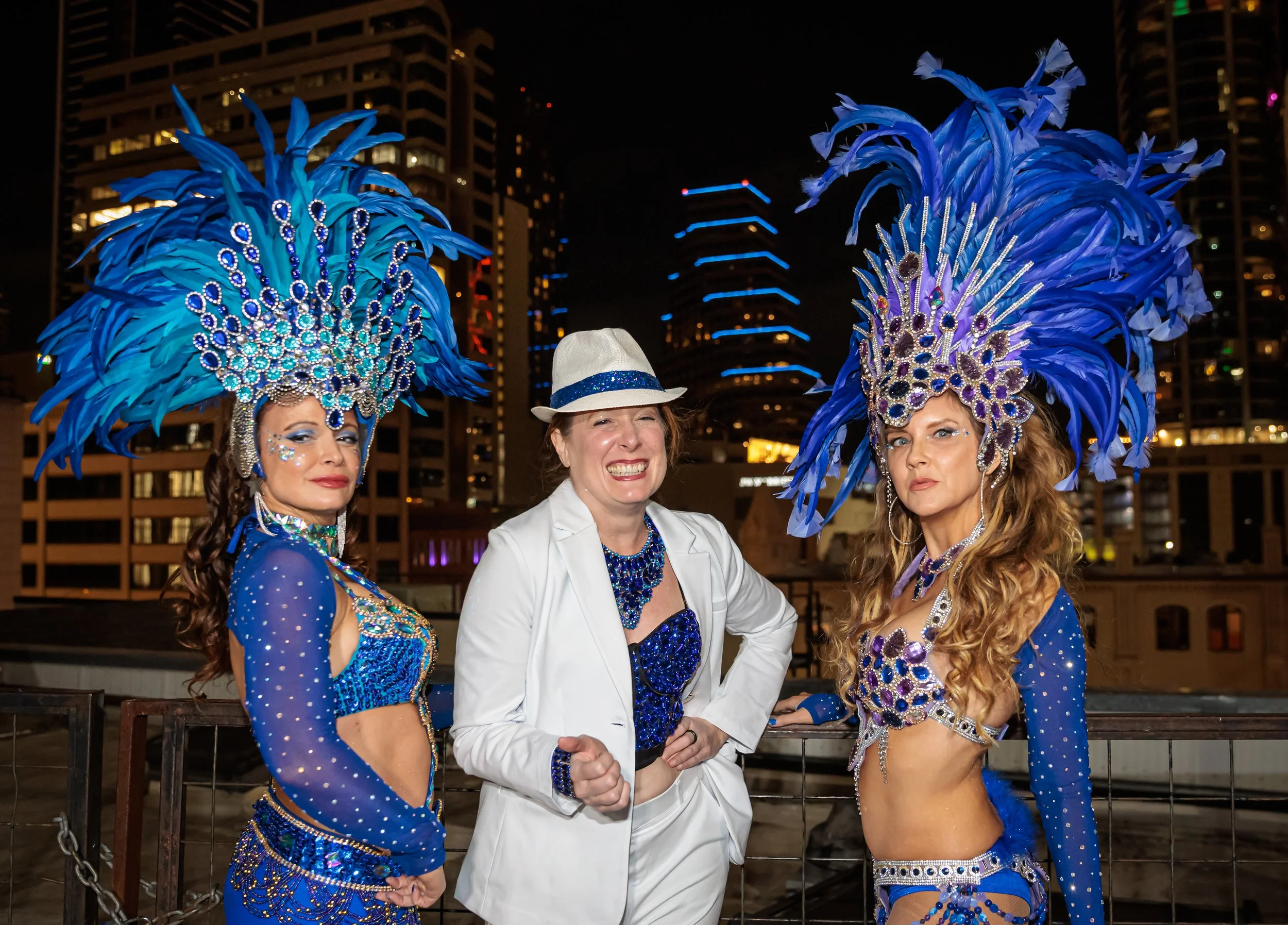 Three women in elaborate blue costumes with feathers and rhinestones, one of whom is smiling broadly in the center, while the other two have flirty expressions. They are standing on a rooftop at Carnaval AUstin with city buildings in the background.