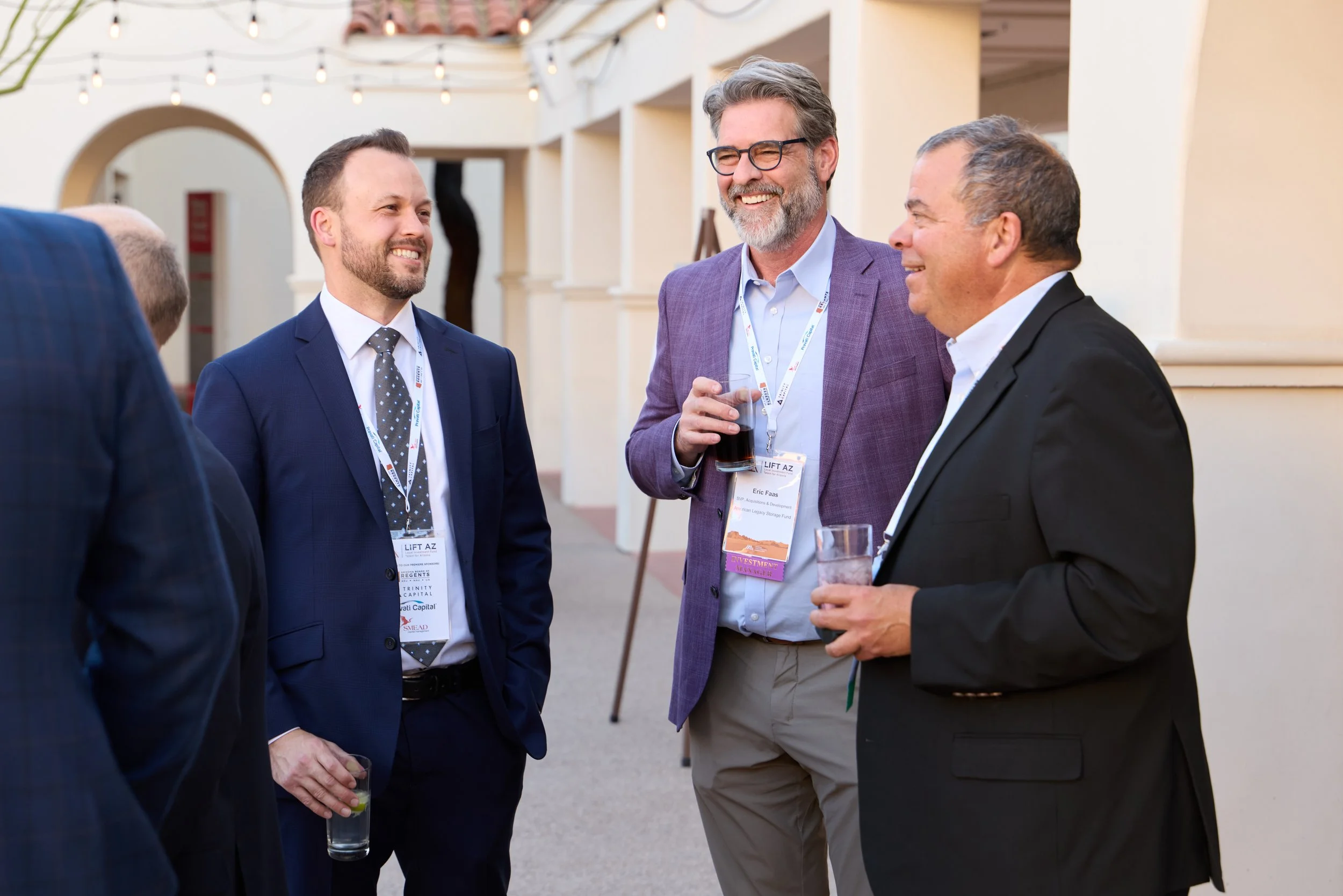 Four men are standing and talking at an outdoor event. They are dressed in business attire, holding drinks, and wearing event badges. The background has string lights and an arched doorway.