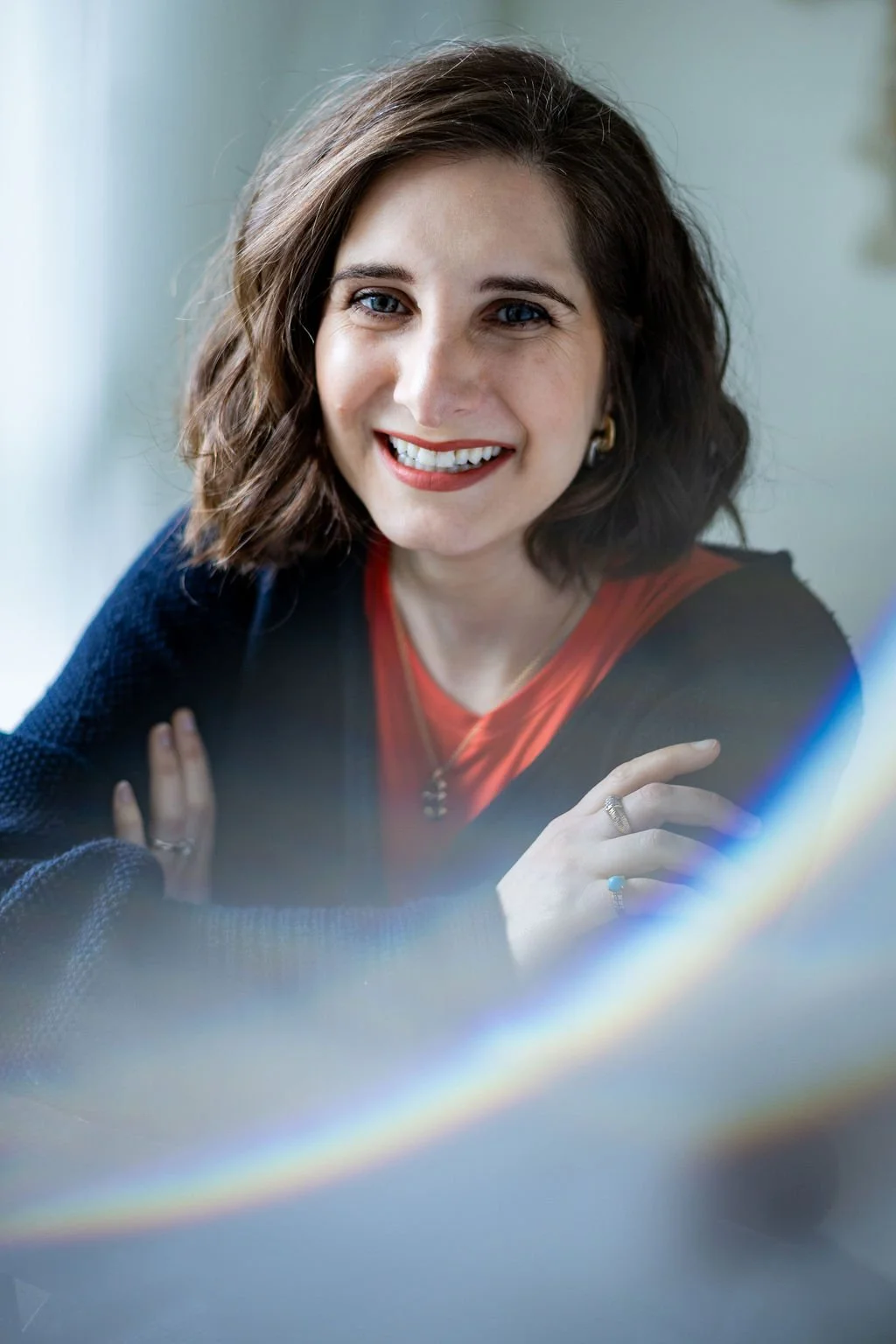 A woman with shoulder-length wavy brown hair, smiling with blue eyes, wearing a red top and black cardigan, sitting with her arms crossed.