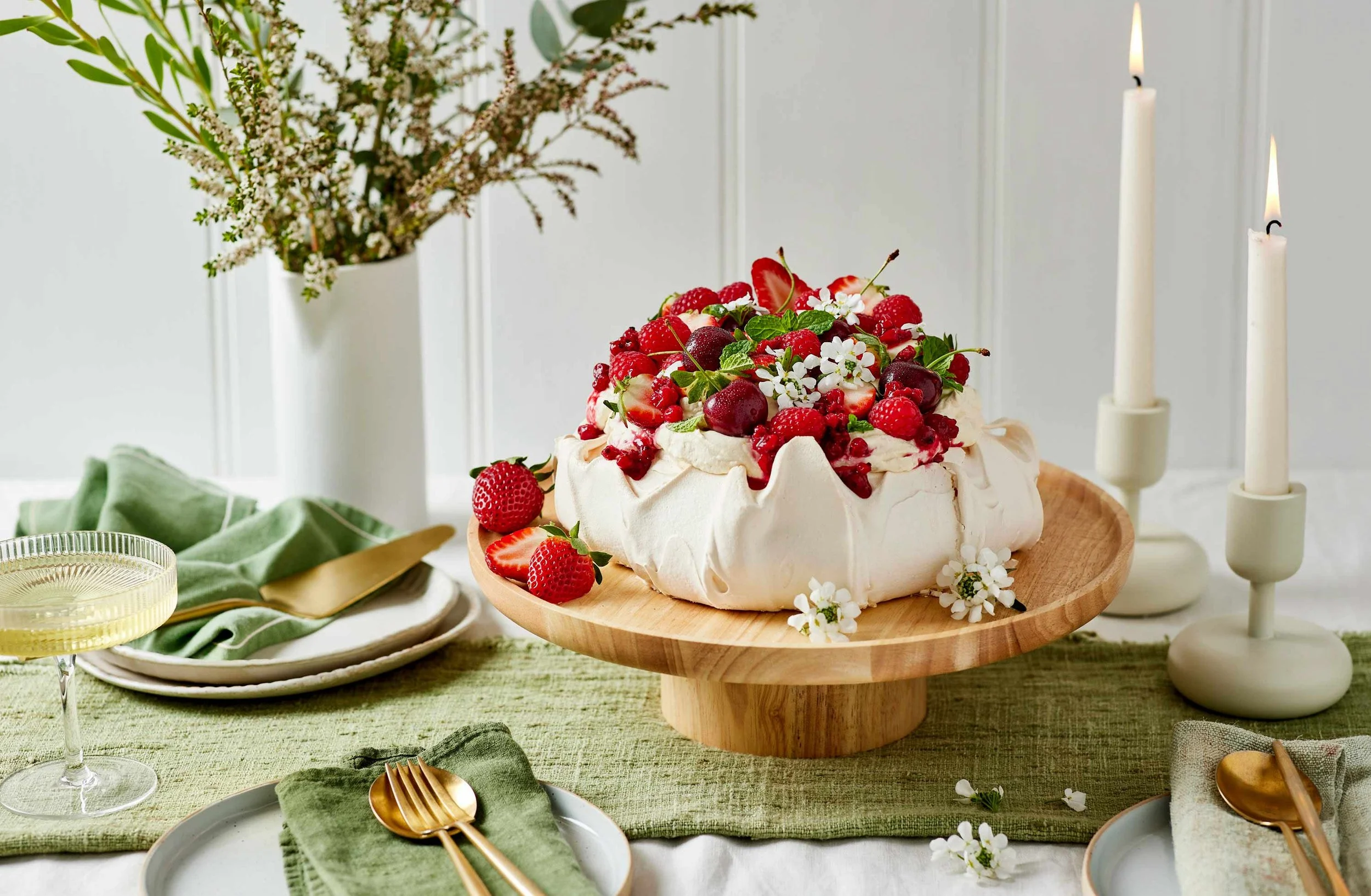 A pavlova cake topped with strawberries, raspberries, cherries, and mint, sitting on a wooden cake stand with a white floral and greenery arrangement and candles in the background.