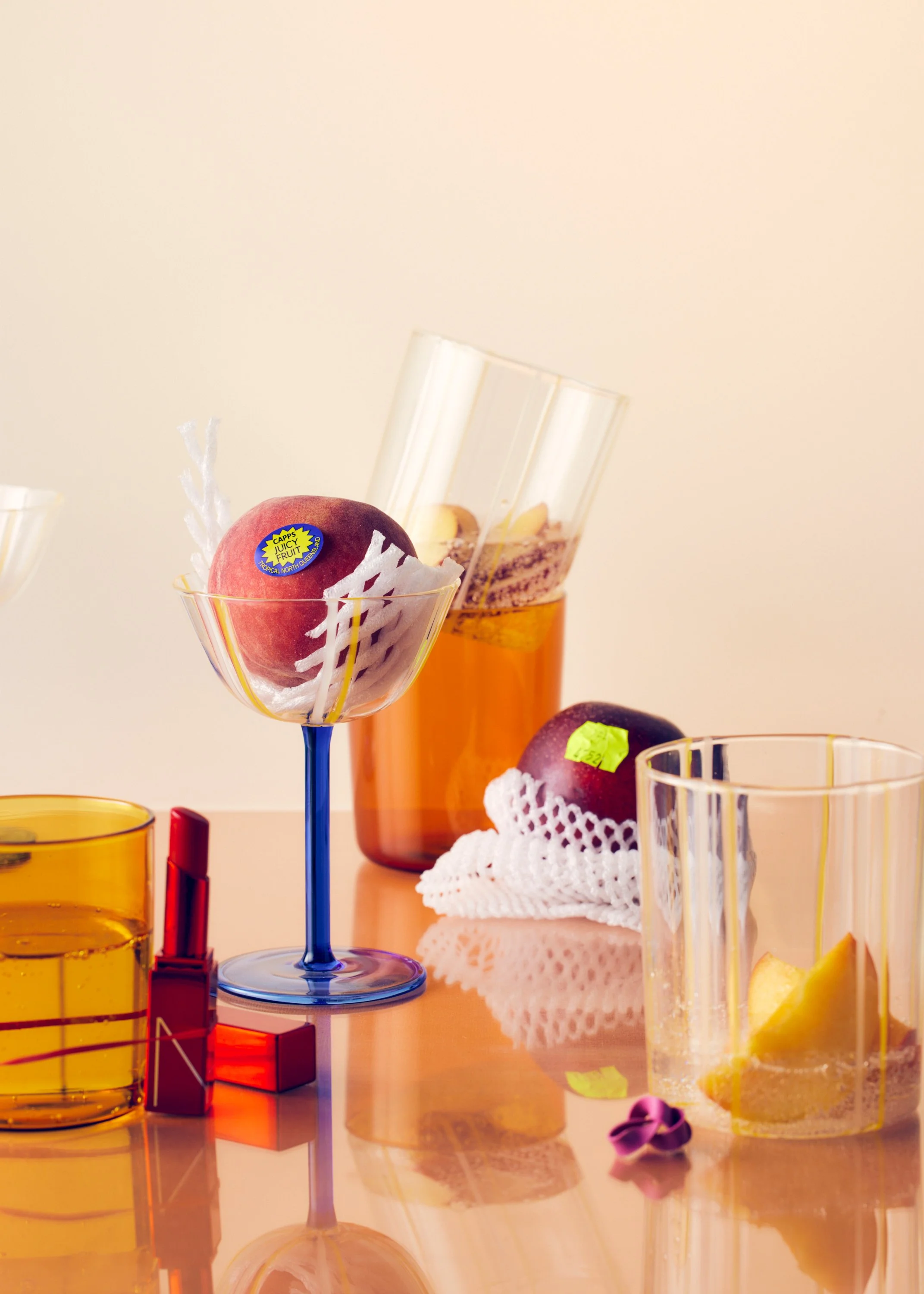 A pink peach with a sticker that reads "Caps Juicy Fruit" sitting in a glass bowl with white decorative paper, placed on a wooden surface. In the background, there is a tall glass with yellow liquid and a straw, a wrapped item, and additional glasses