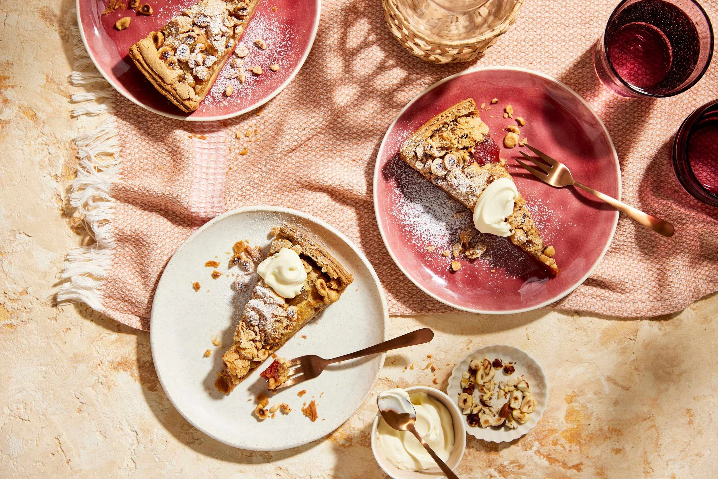 Various slices of dessert pies topped with powdered sugar and nuts on pink and white plates, accompanied by small bowls of whipped cream and glasses of red wine on a textured pink fabric tablecloth.