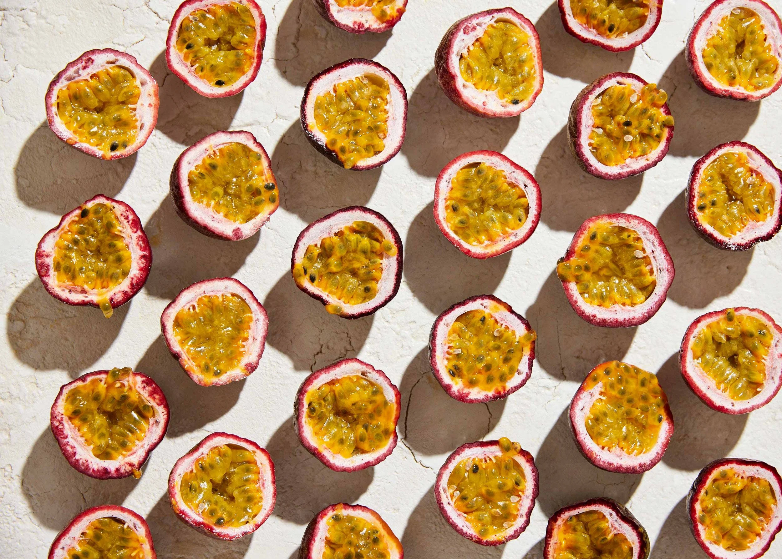 A flat lay of halved passion fruits with bright yellow pulp and black seeds on a light-colored surface, casting shadows.
