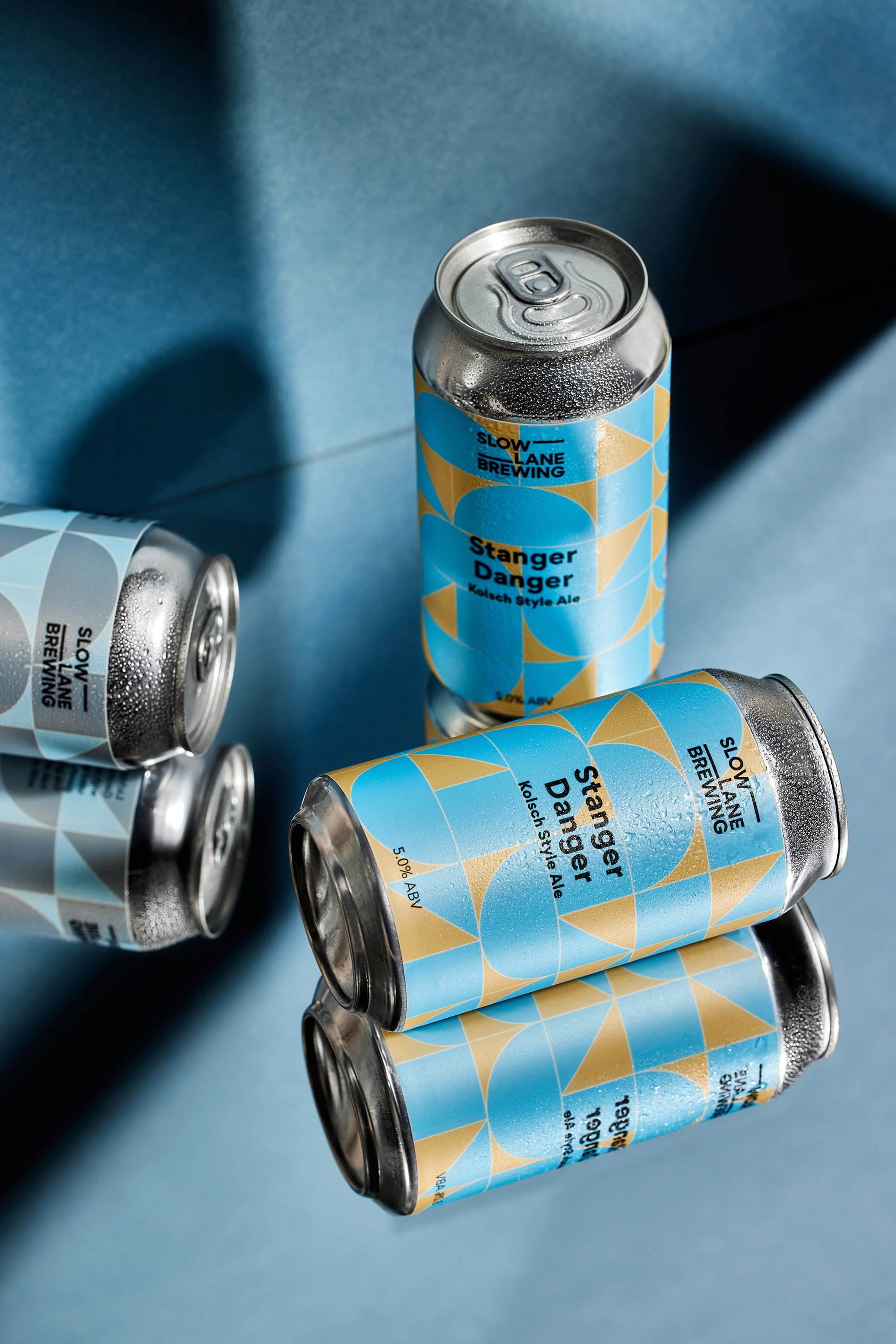 Cans of Slow Lane Brewing Stanger Danger Kolsch Style Ale beer with geometric yellow and blue label, some cans stacked and some lying on their sides, on a blue background.