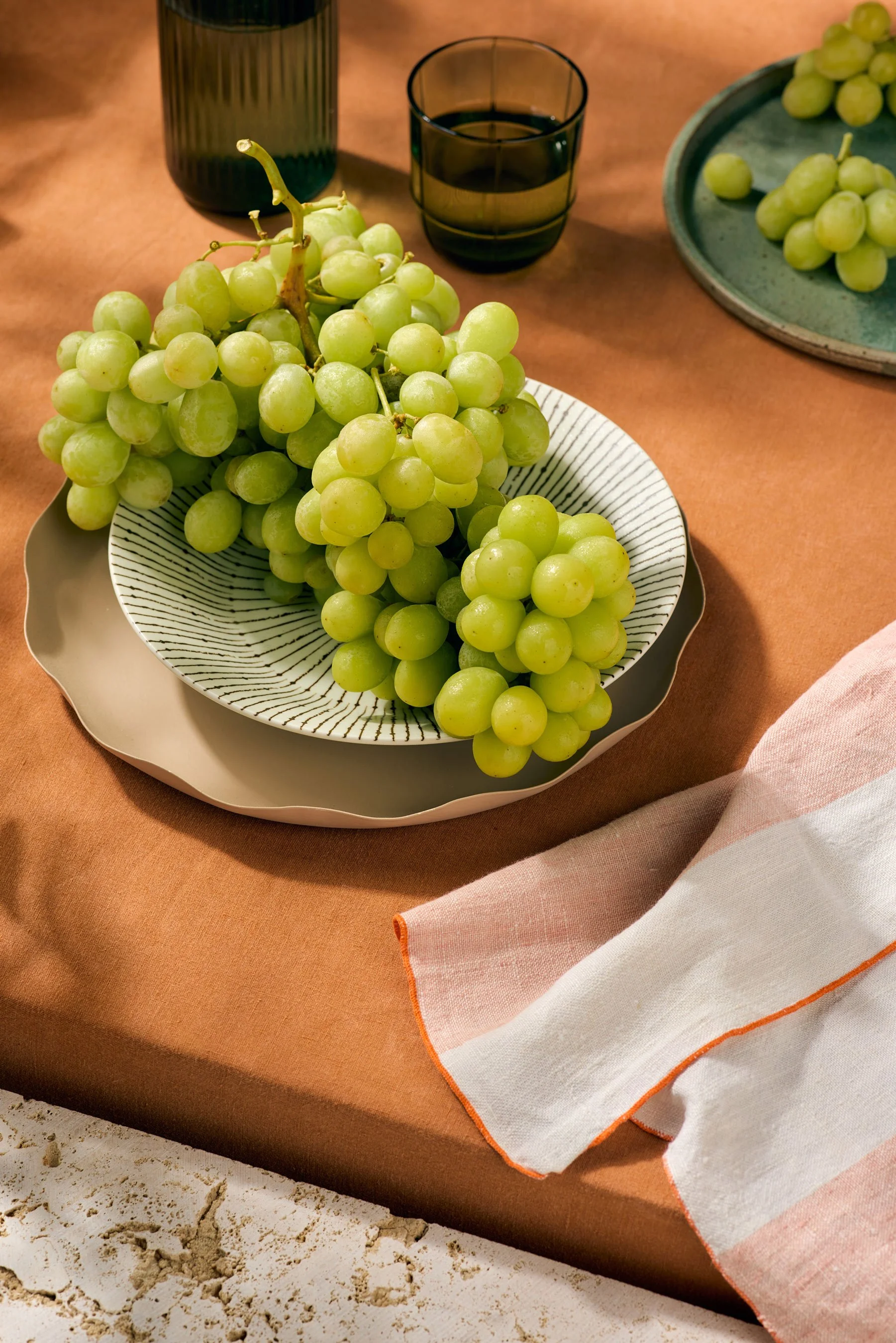 Green grapes on a plate with dark glasses and a bottle on a brown table, with a linen napkin with orange trim beside the table.