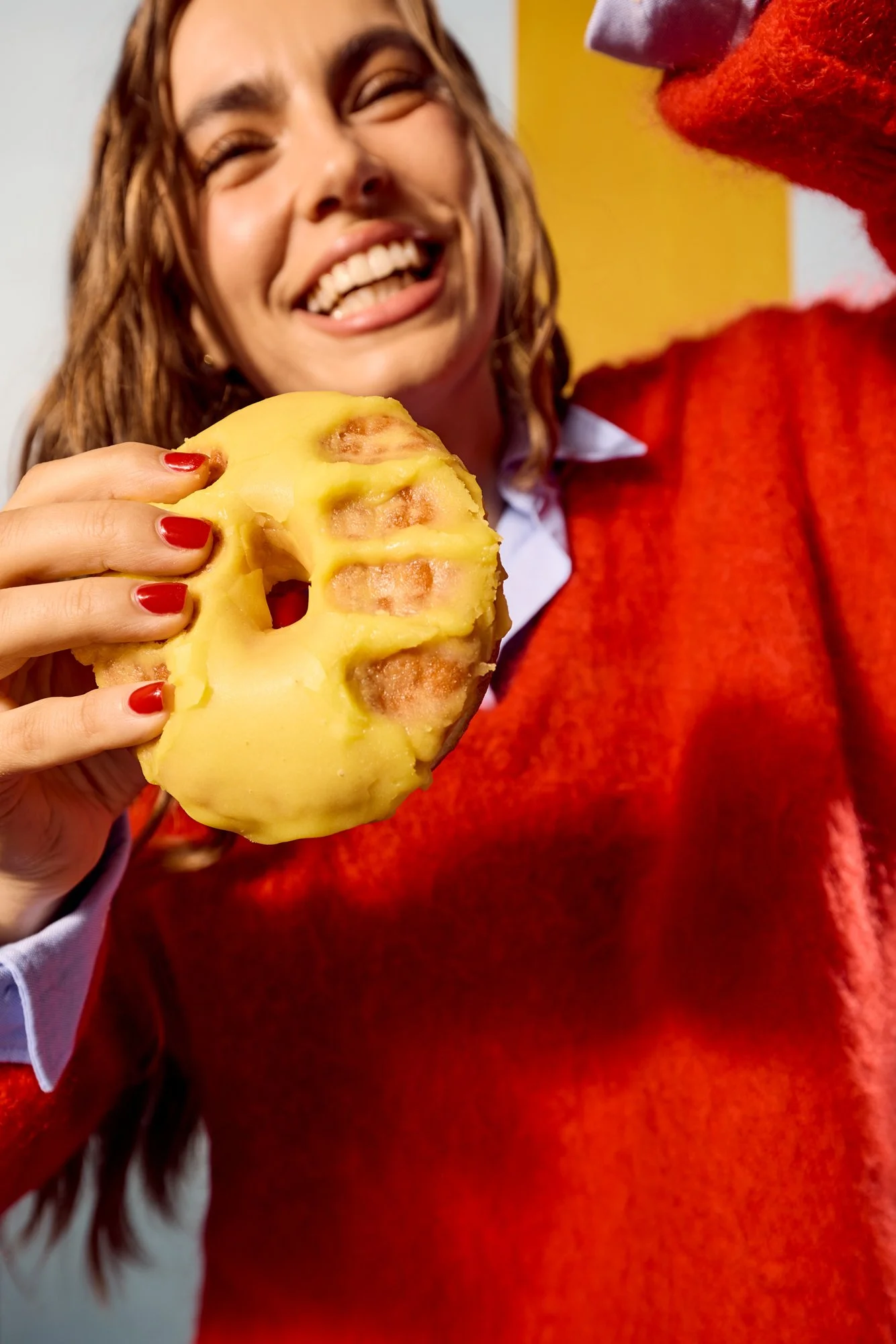 A woman with red nails and long brown hair is smiling as she holds a peeled pineapple in her hand. She is wearing a red sweater and a white shirt underneath. A person next to her is wearing a red and purple costume or glove, possibly holding or offer
