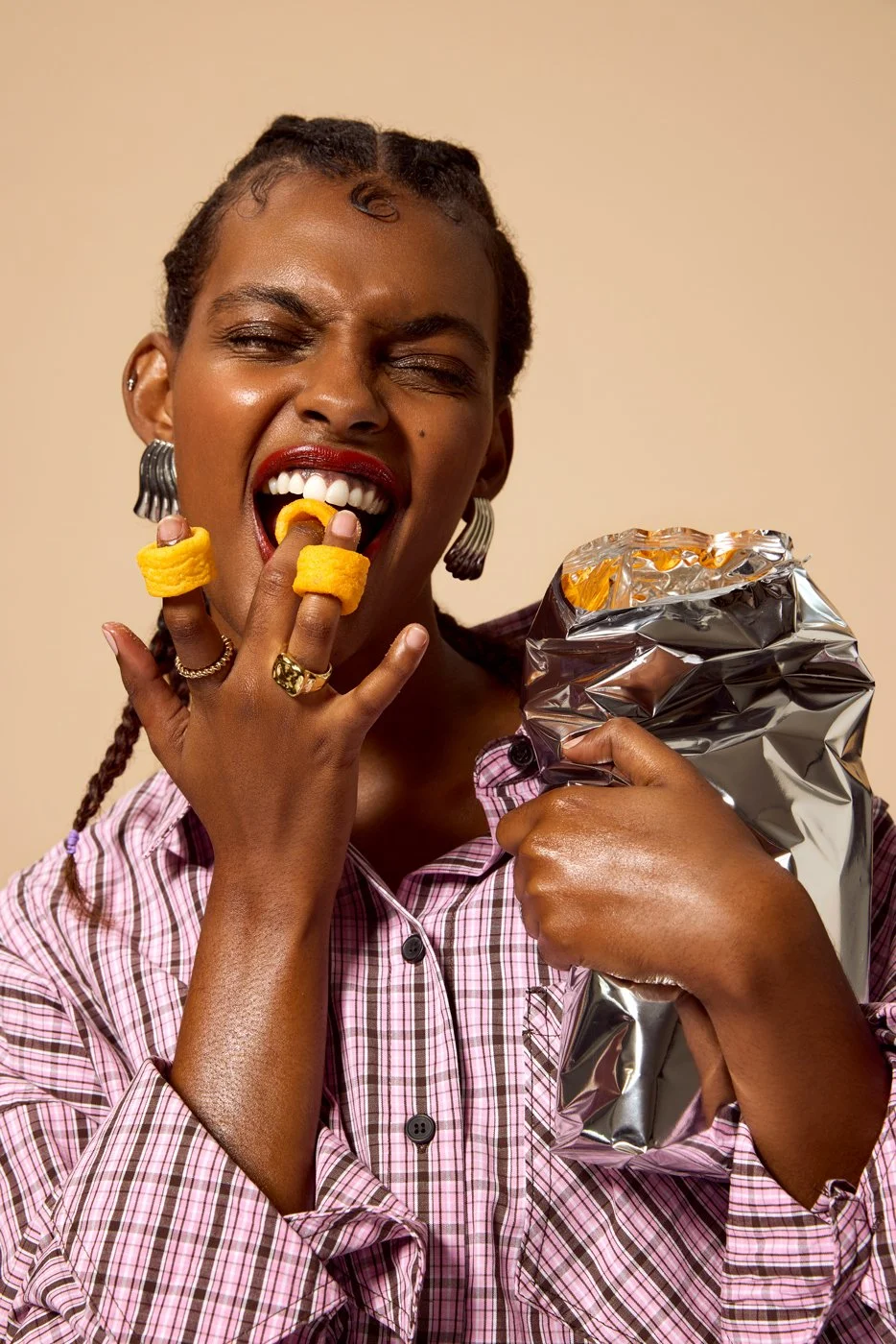 A woman with braided hair and earrings eating potato chips from a silver bag.