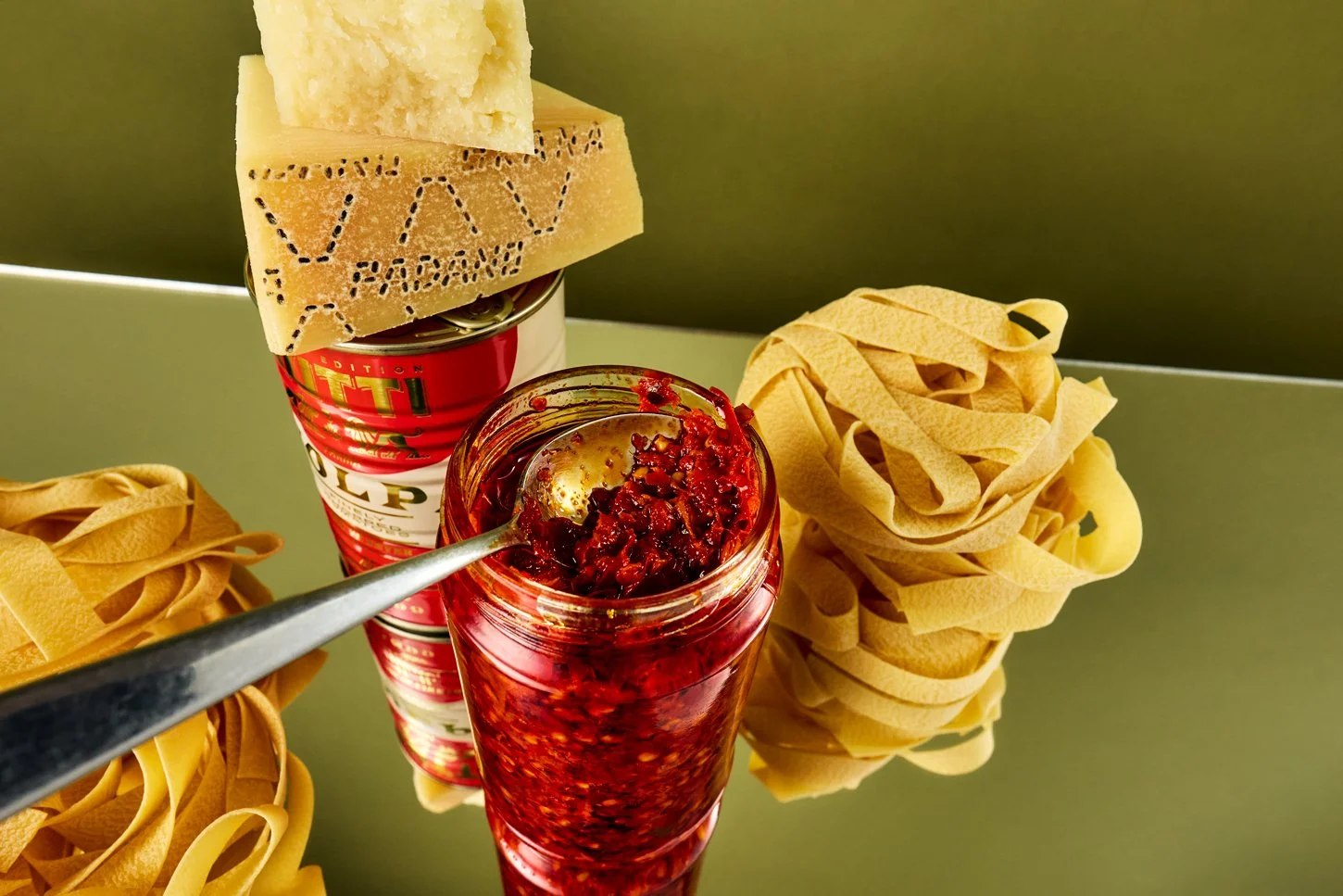 Open jar of red chili pepper sauce with a spoon, Mutti can of diced tomatoes, parmesan cheese block balanced on top, uncooked fettuccine pasta bundles on a reflective surface.