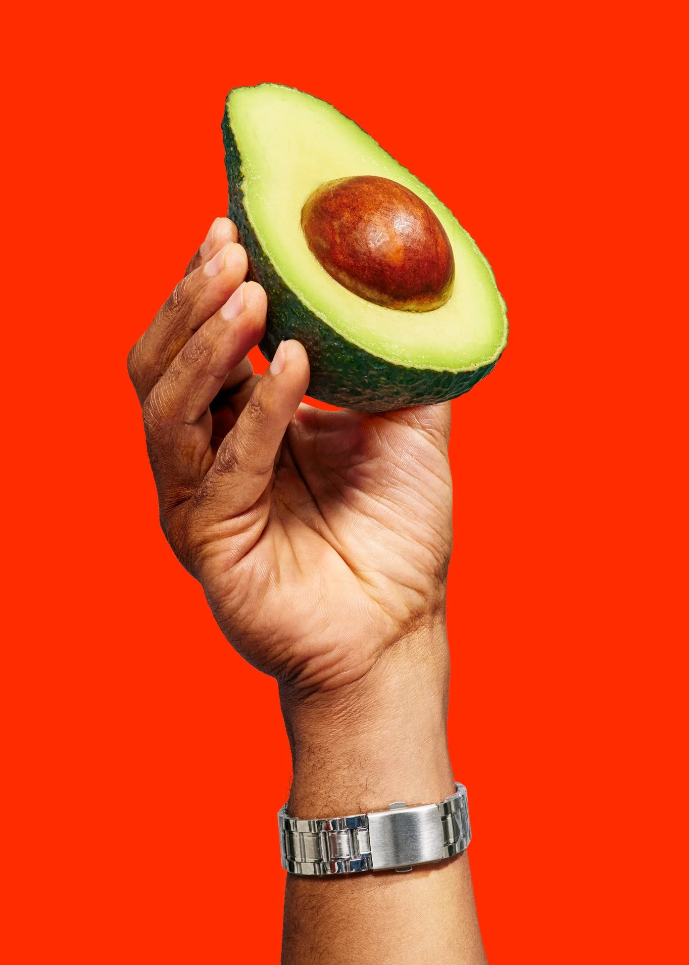 Doordash advertising campaign ''smash it''.  Hand holding half an avocado with seed, silver watch on wrist, red background.