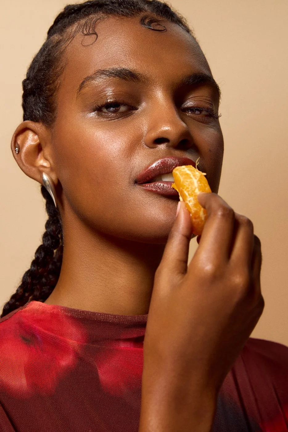 Close-up of a black woman of colour with braided hair, glossy skin, holding a mandarin about to eat it, wearing a red top and earrings against a beige background.