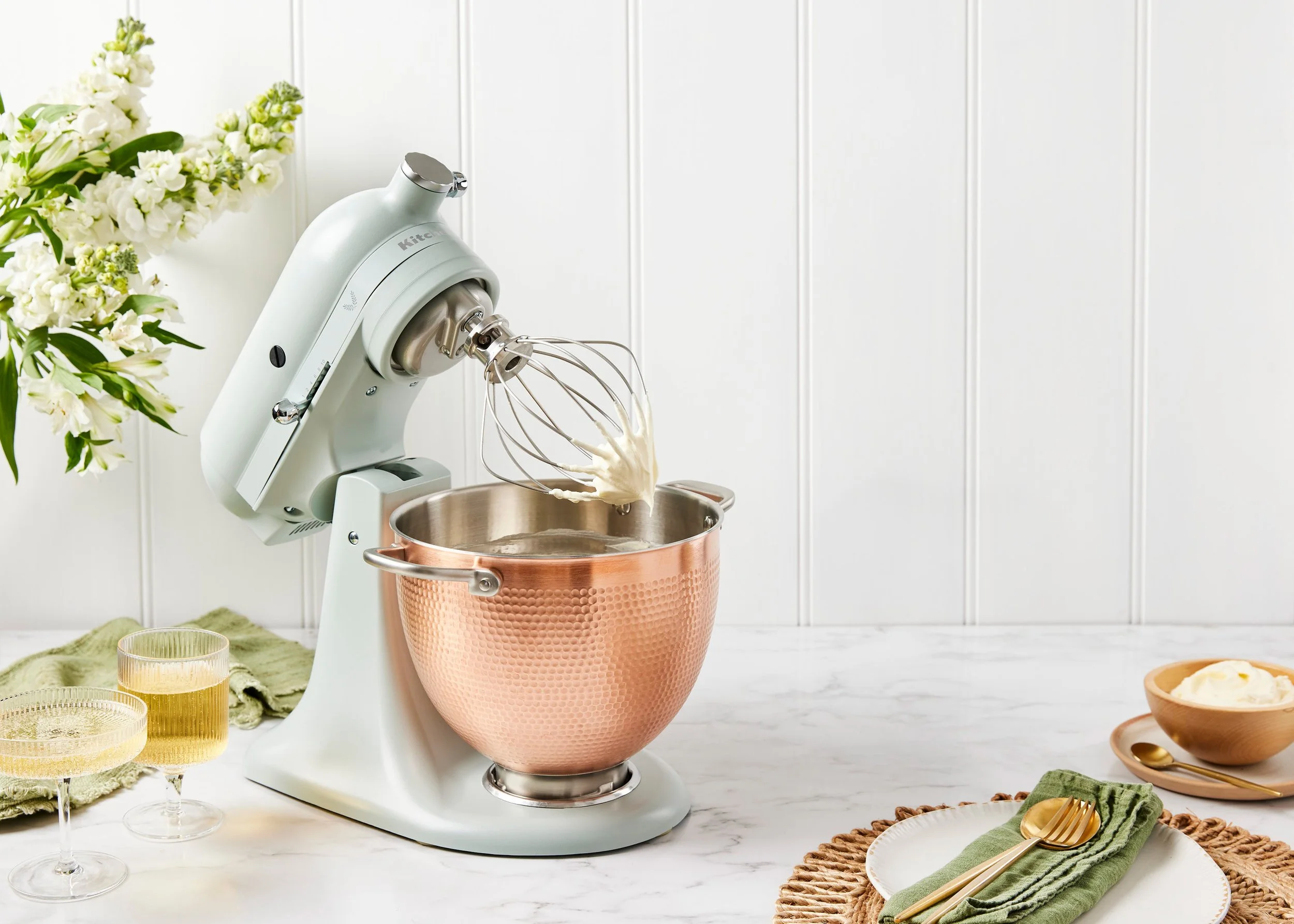 A kitchen stand mixer with a copper bowl, whisks attached, on a white marble countertop. To the left, there are three glasses filled with a yellow beverage, and behind them, a bouquet of white flowers. To the right, a small bowl with a pat of butter 