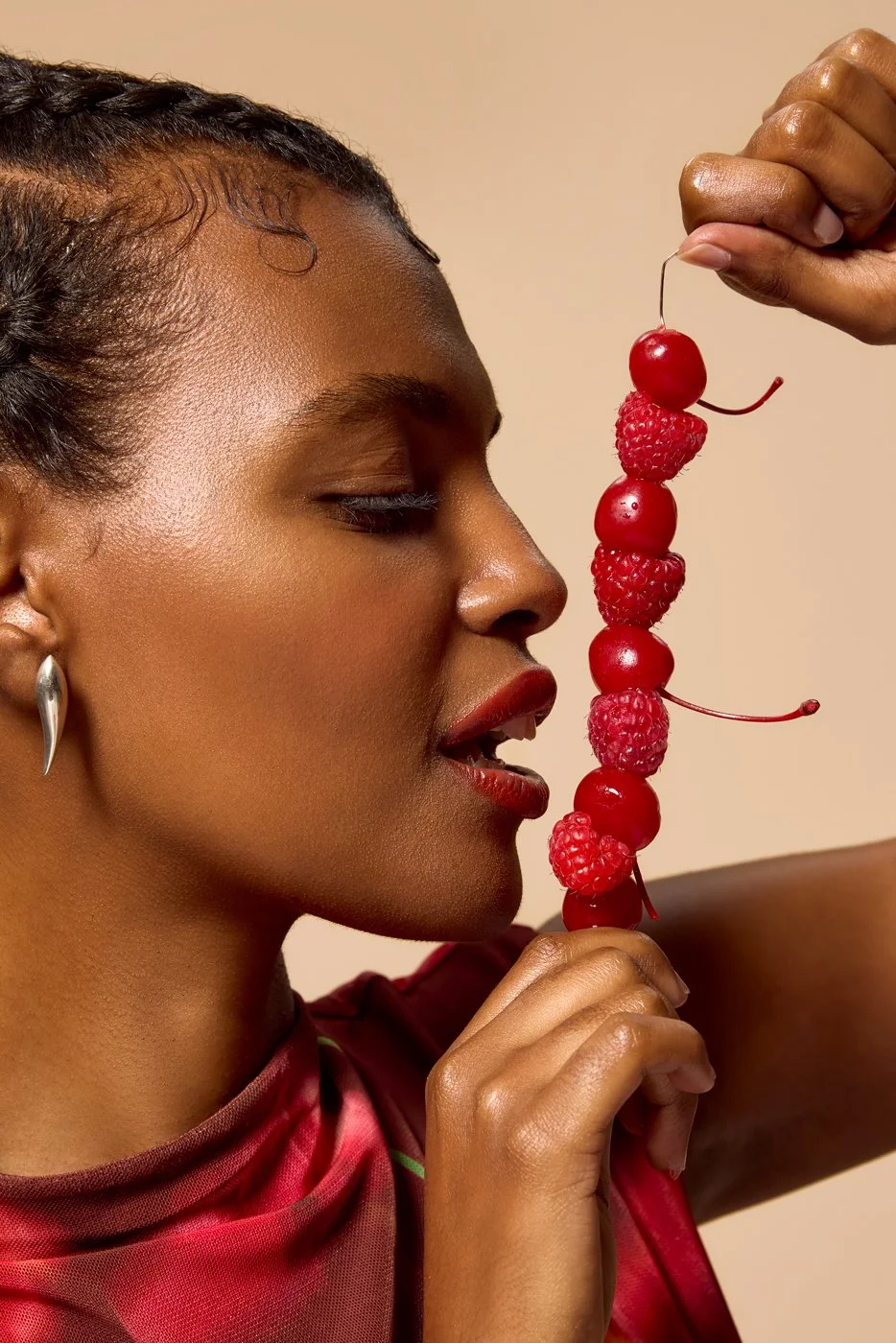 Close-up of a woman with short curly hair, wearing earrings and red lipstick, holding a skewer of alternating raspberries and cherries near her face.