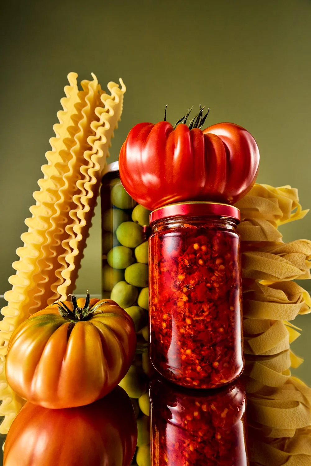 Assorted heirloom tomatoes, jar of chili sauce, green olives and uncooked pasta on a reflective surface.
