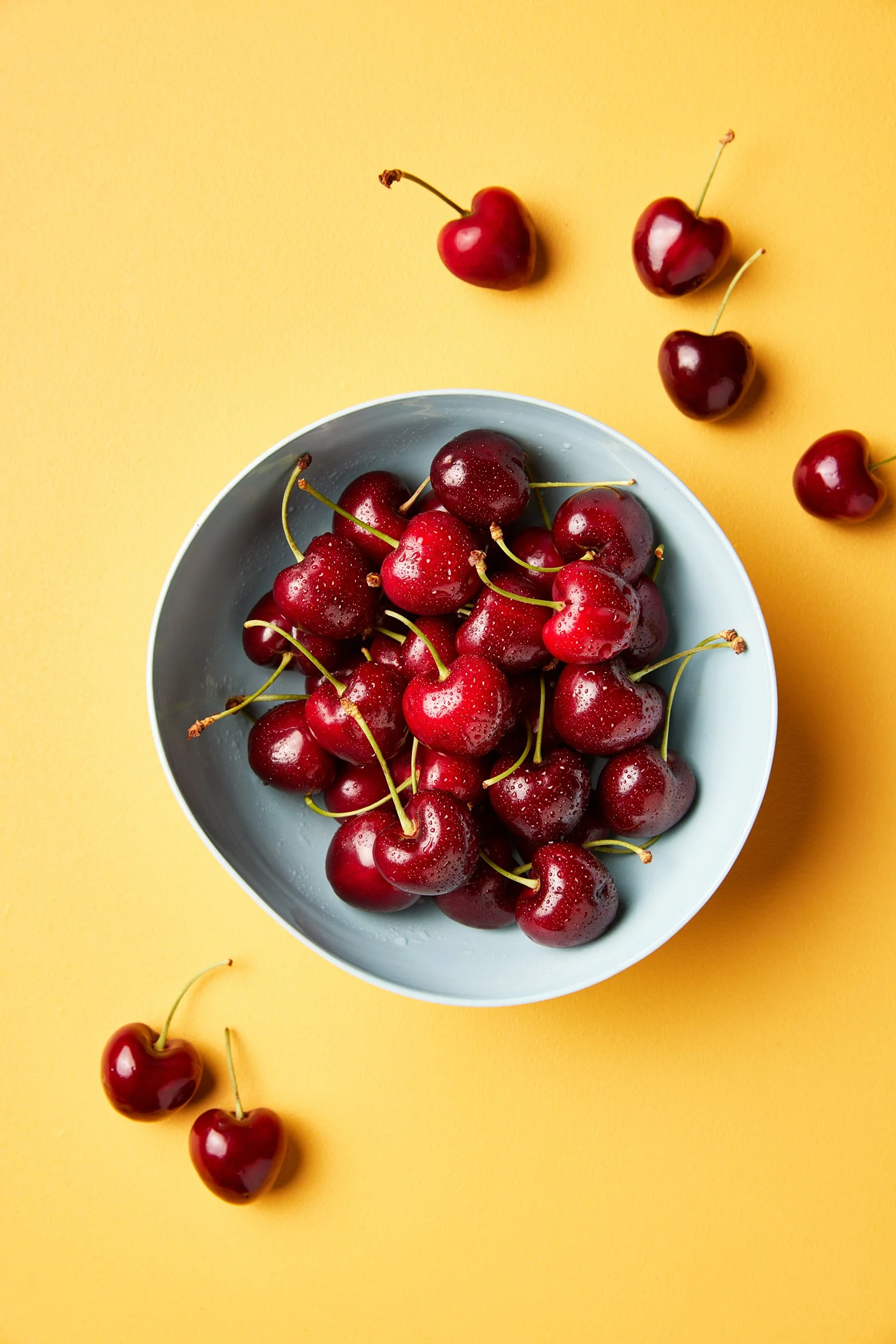 A bowl of fresh red cherries on a yellow background, with some cherries spilling outside the bowl.