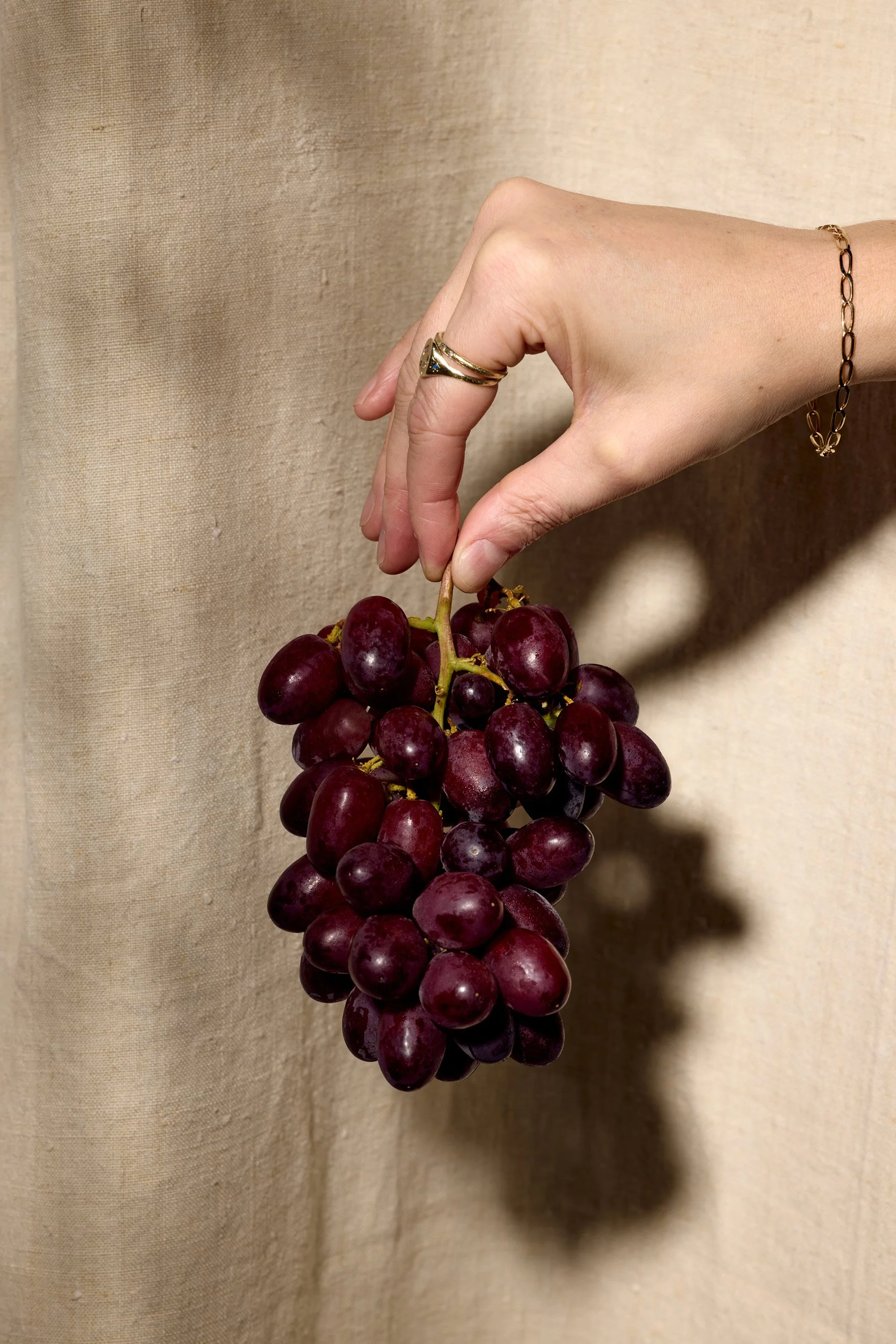 A hand wearing rings and a bracelet holding a bunch of red grapes against a beige fabric background.