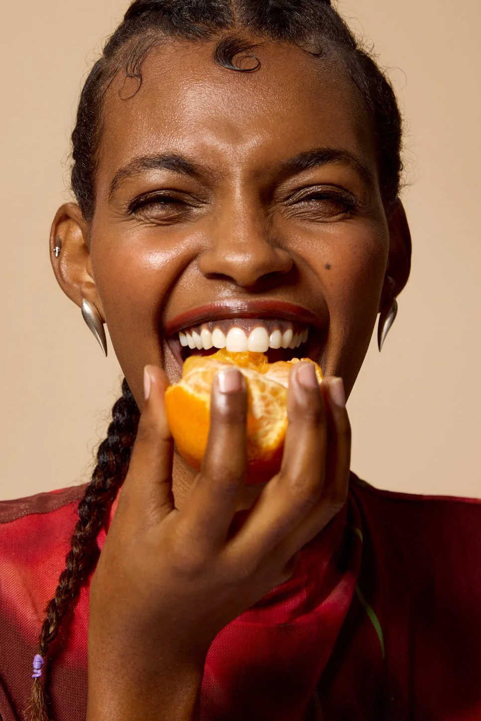 A black woman of colour with glossy skin and braided hair, wearing earrings and a red top, is smiling while holding an orange and taking a bite of it.