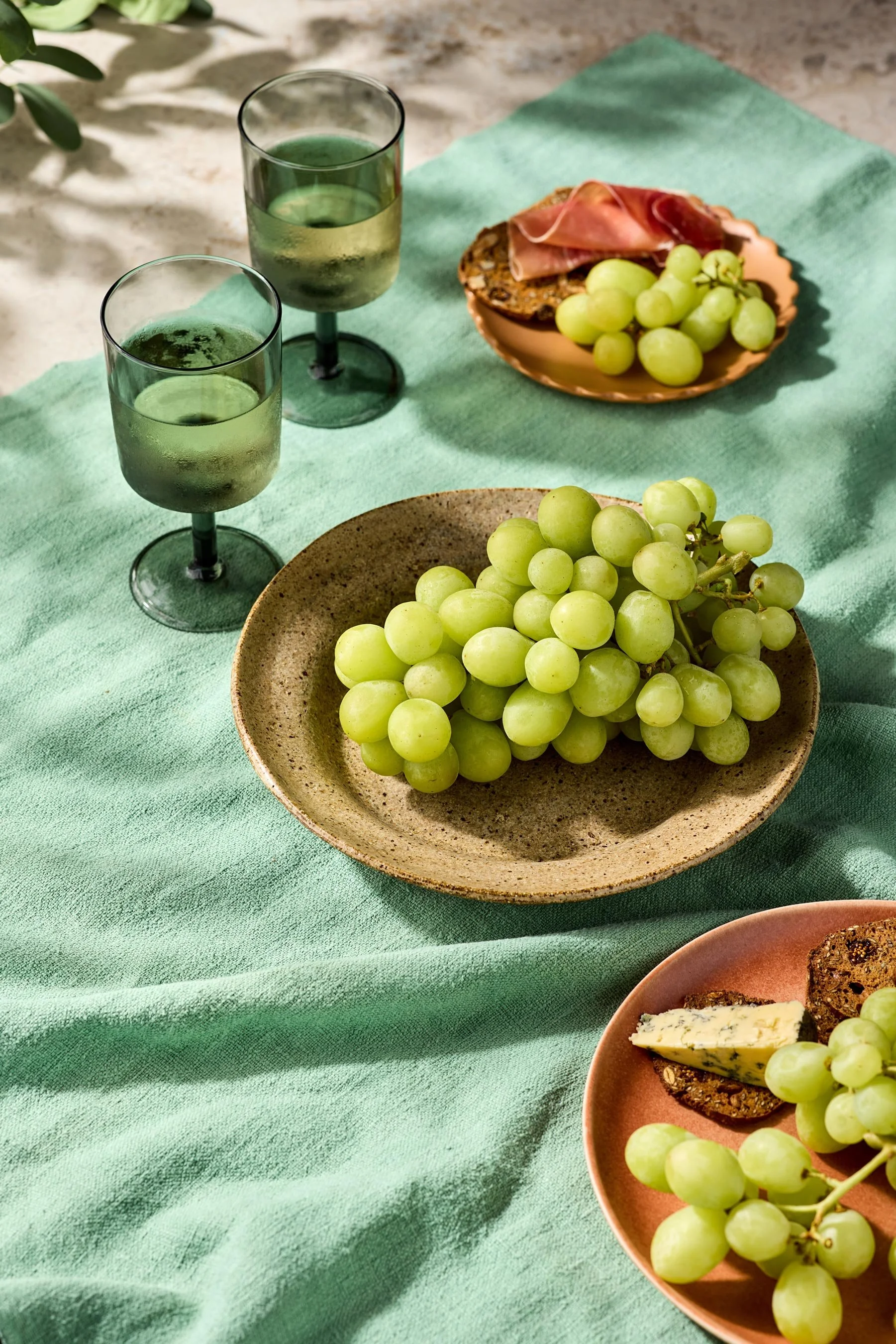 Two glasses of white wine, a plate of green grapes, and a plate with slices of cheese and crackers on a green tablecloth.