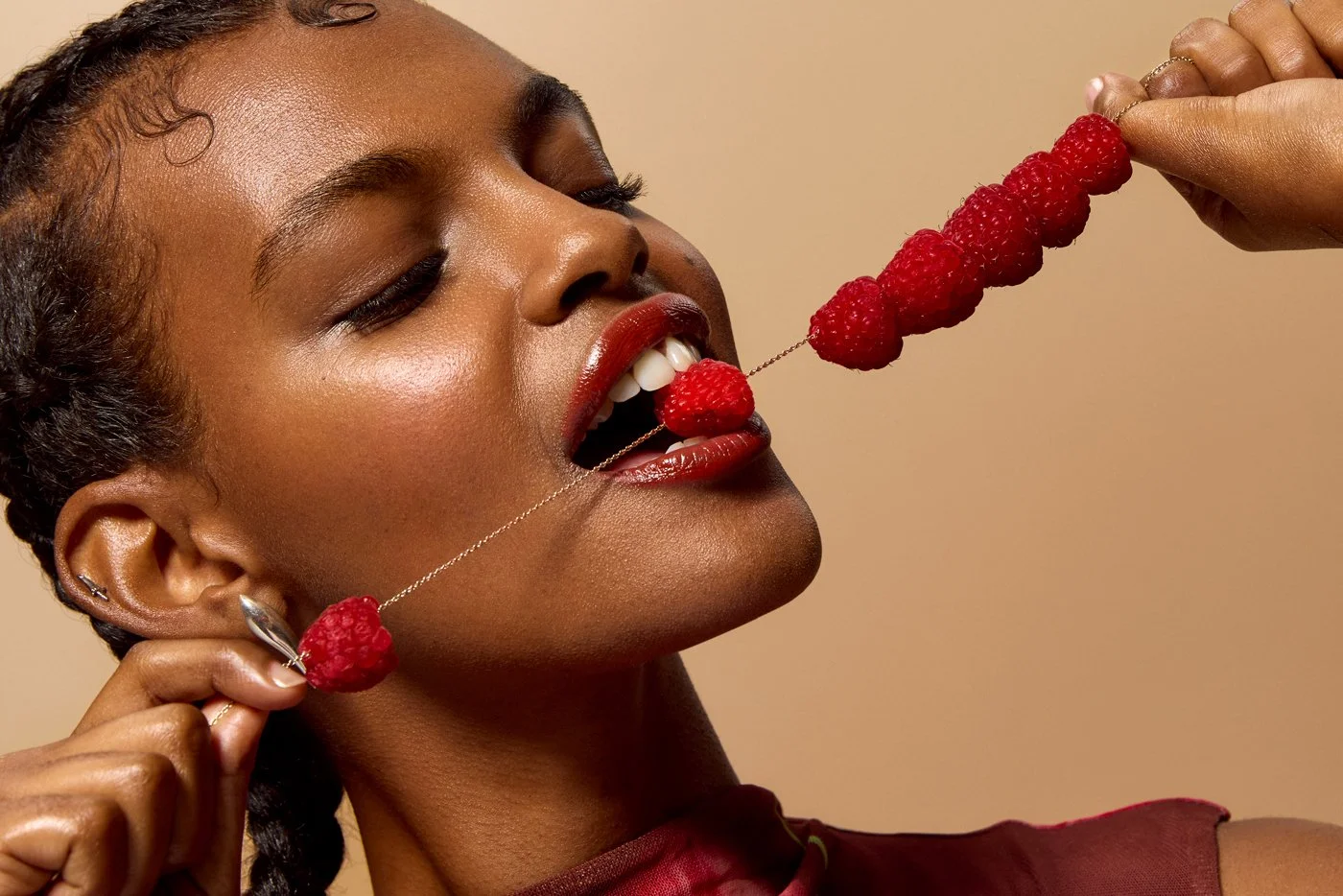 A black woman of colour with braided hair, red lipstick, and earrings is about to eat a raspberry necklace, which is held by another hand, glossy beauty lighting on the skin