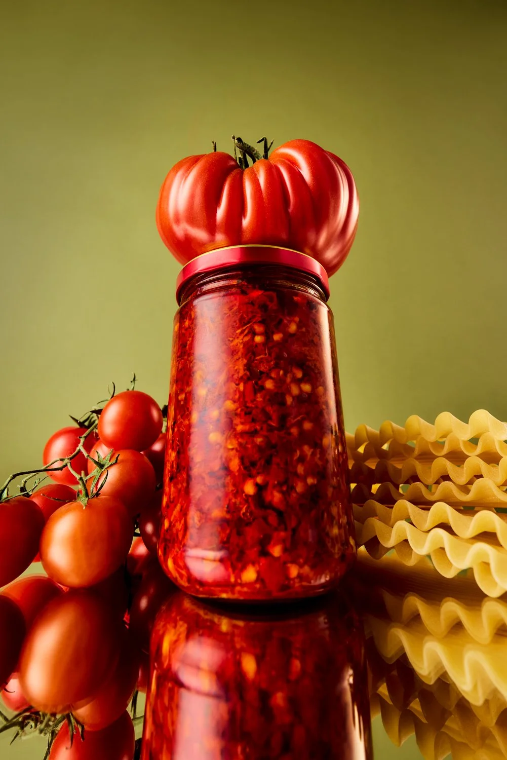 A jar of spicy red chili sauce with a large heirloom tomato on top, nearby cherry tomatoes on a vine, and uncooked yellow pasta ribbons on the side, all reflected on a glossy surface against a green background.