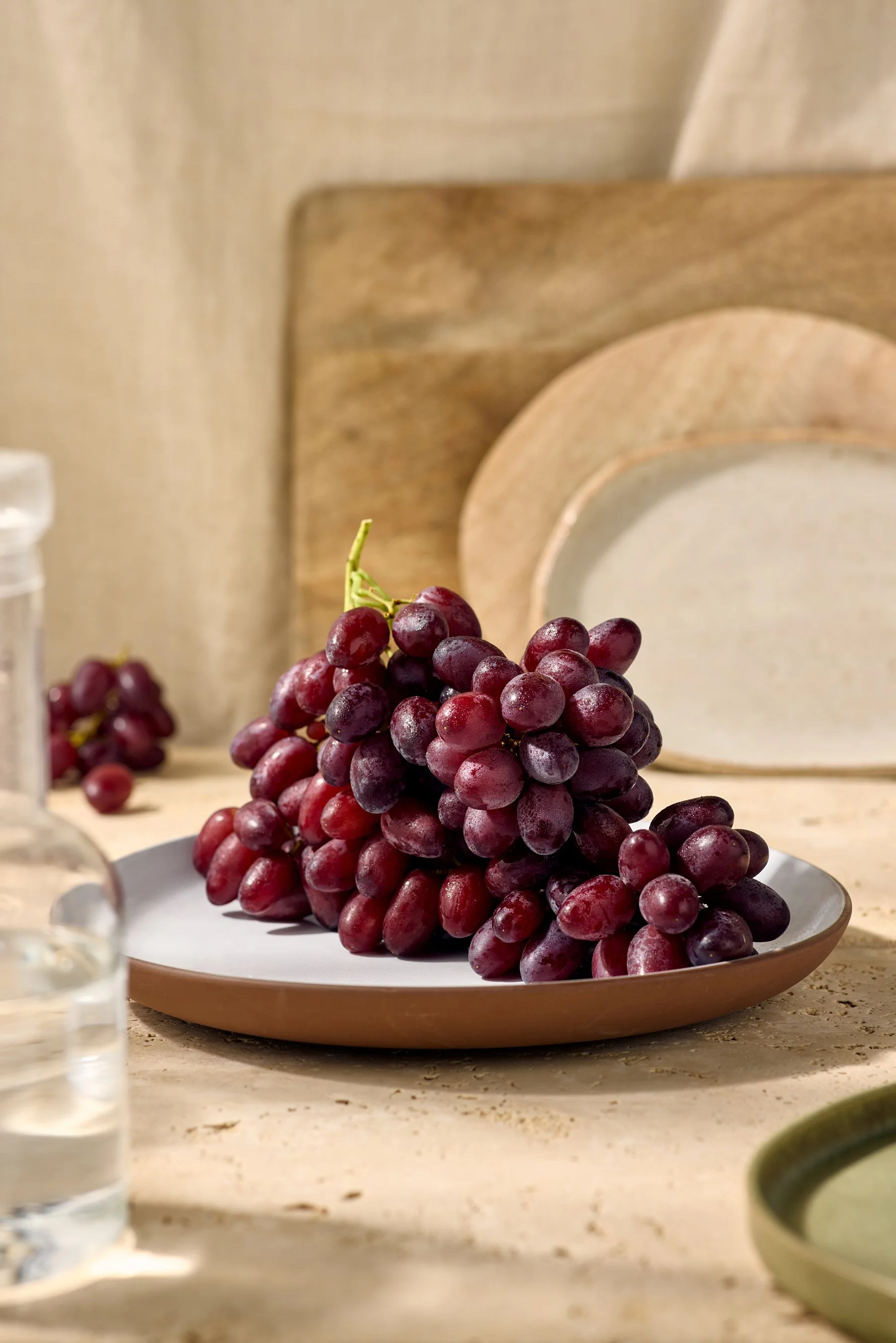 A bunch of red grapes on a white plate on a kitchen countertop with a beige backsplash, a wooden cutting board, a glass of water, and a small bowl of green dip or sauce.