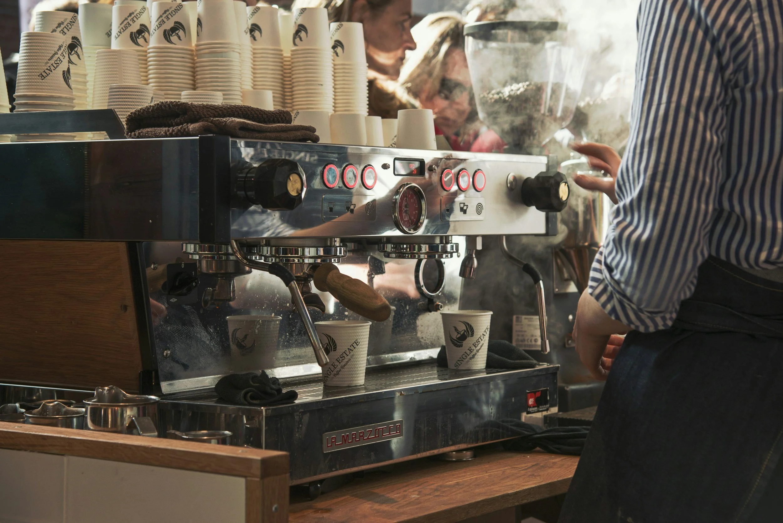 Barista preparing coffee on a commercial espresso machine with steam rising, stacked disposable coffee cups on top, and customers in the background.