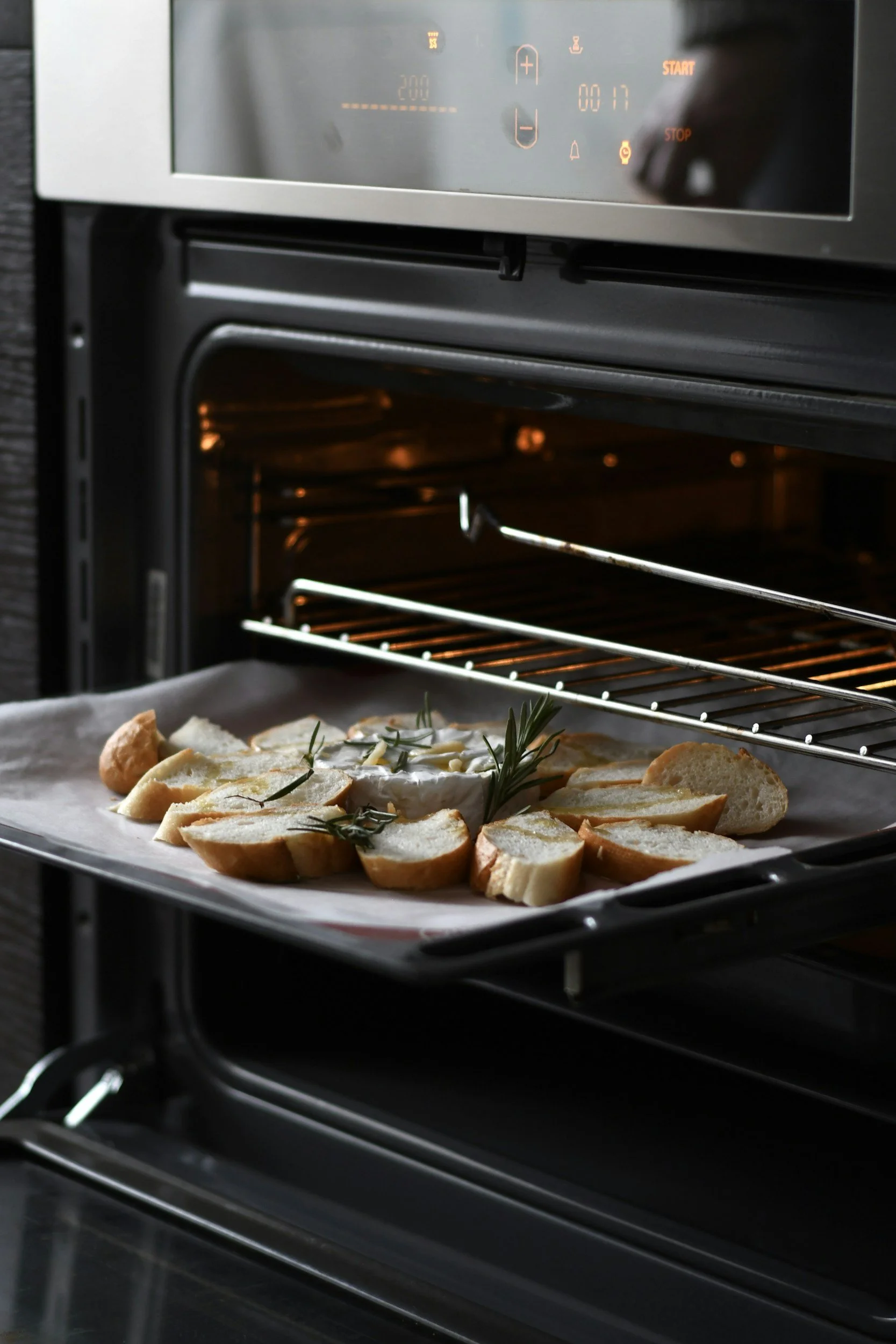 A tray of sliced bread topped with herbs and a small round cheese inside a modern oven with a digital display.