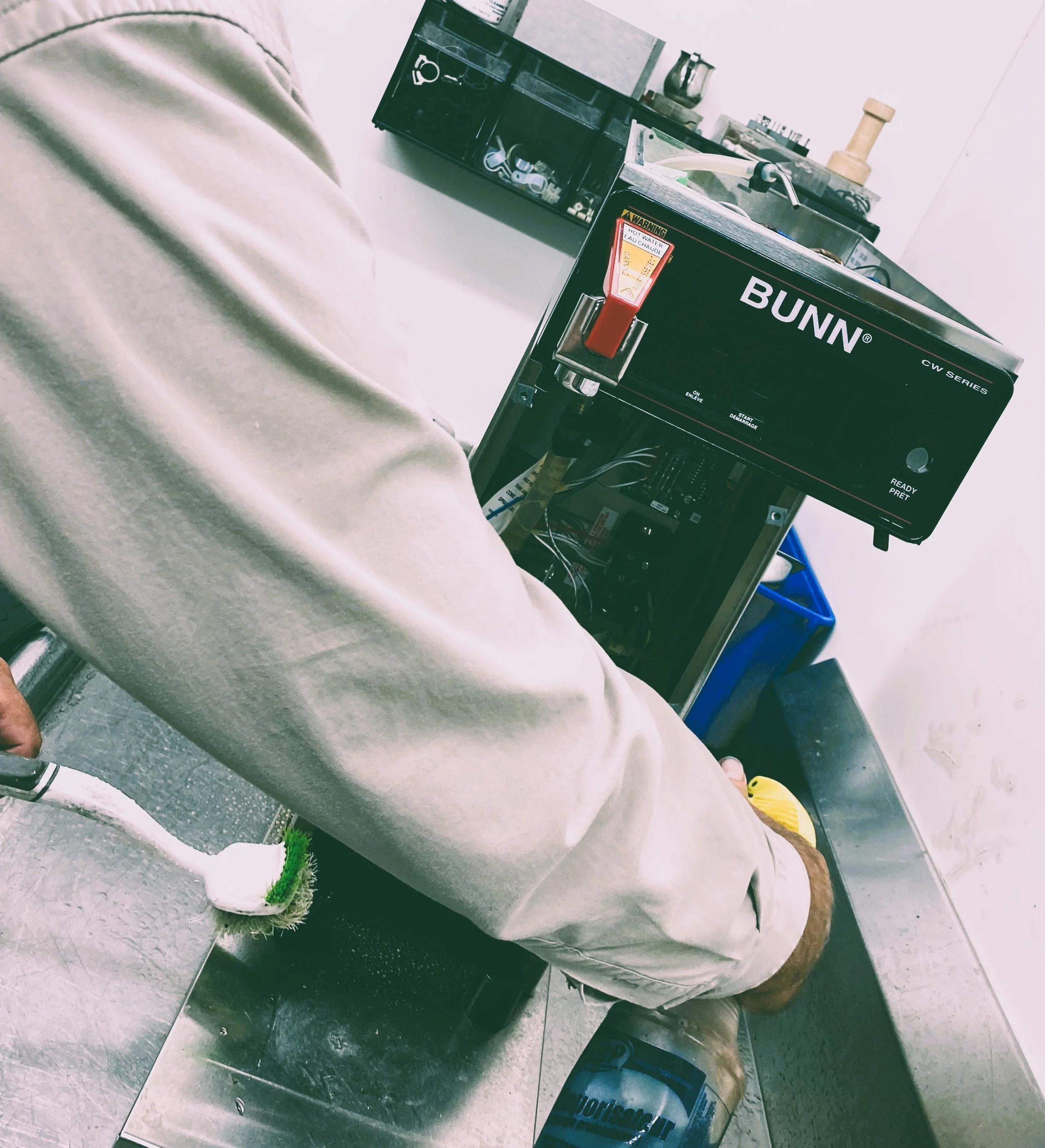 Technician working on a Bunn coffee machine in a commercial kitchen, with various tools and equipment in the background.