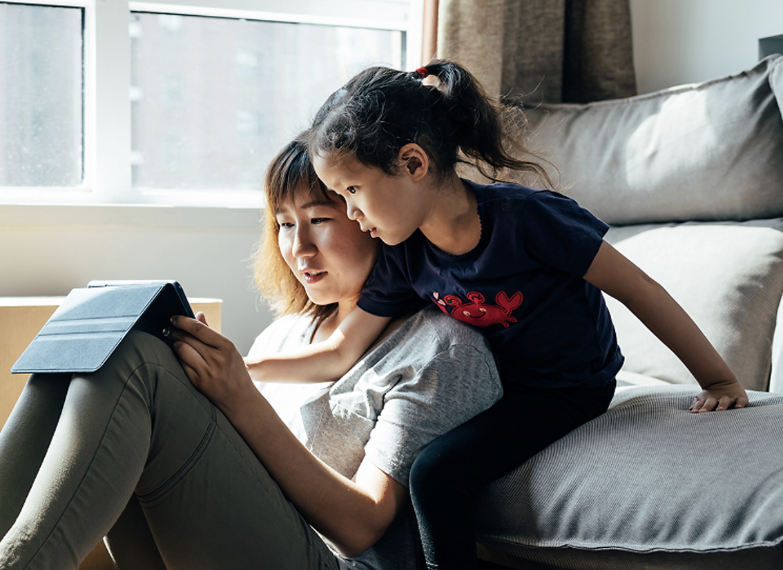 A woman and girl sit near a couch watching a tablet