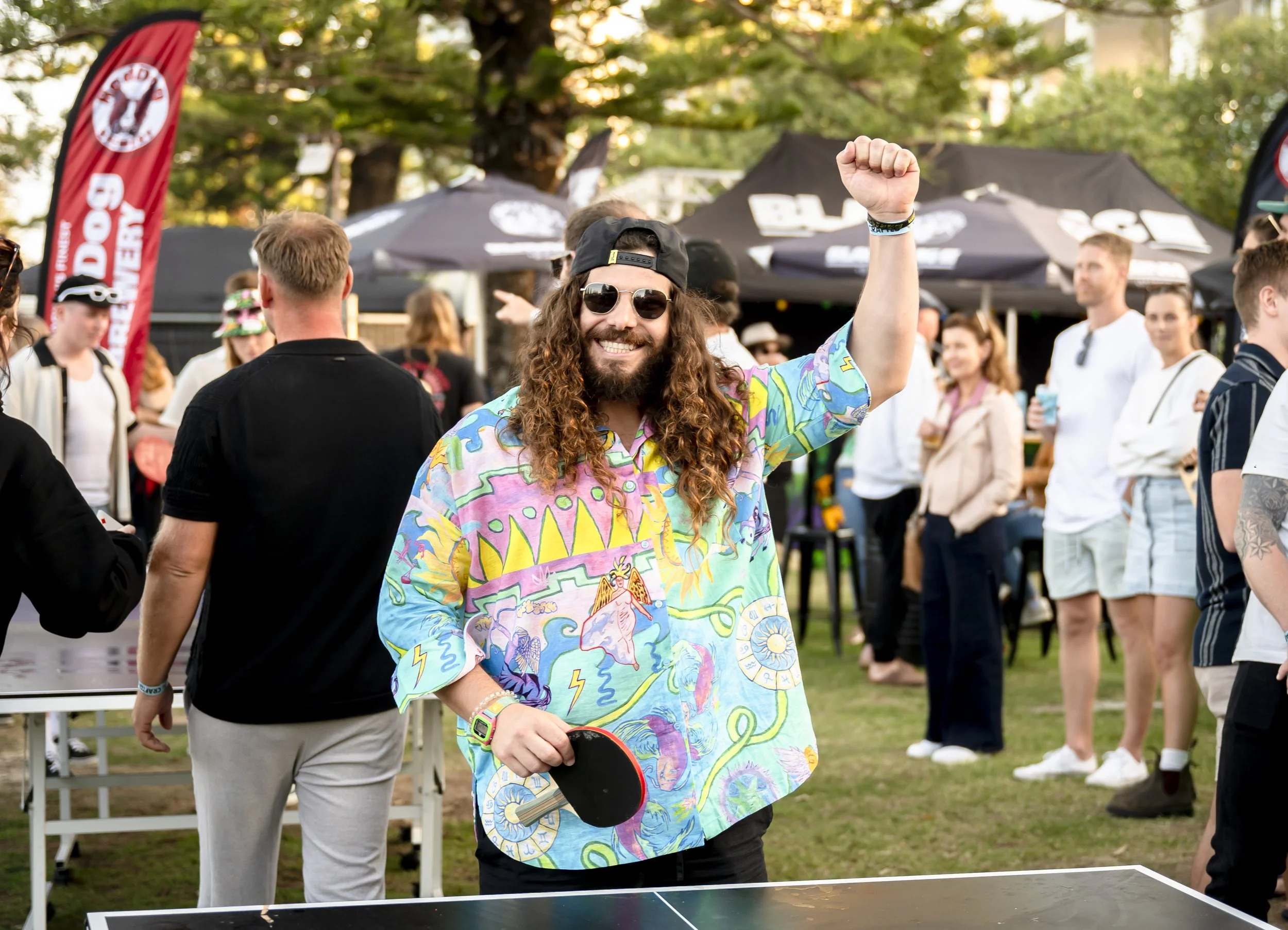 A man with long curly hair, wearing sunglasses, a colorful patterned shirt, and a backwards cap, is smiling and raising his fist in celebration at an outdoor event with many people, tents, and trees in the background.