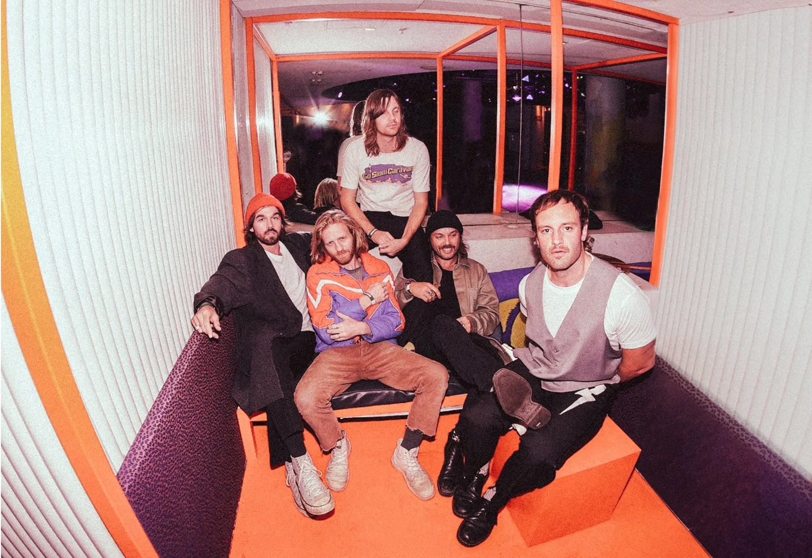 Five men sitting and standing inside a small booth with orange framing, with a neon-lit stage visible outside the booth.