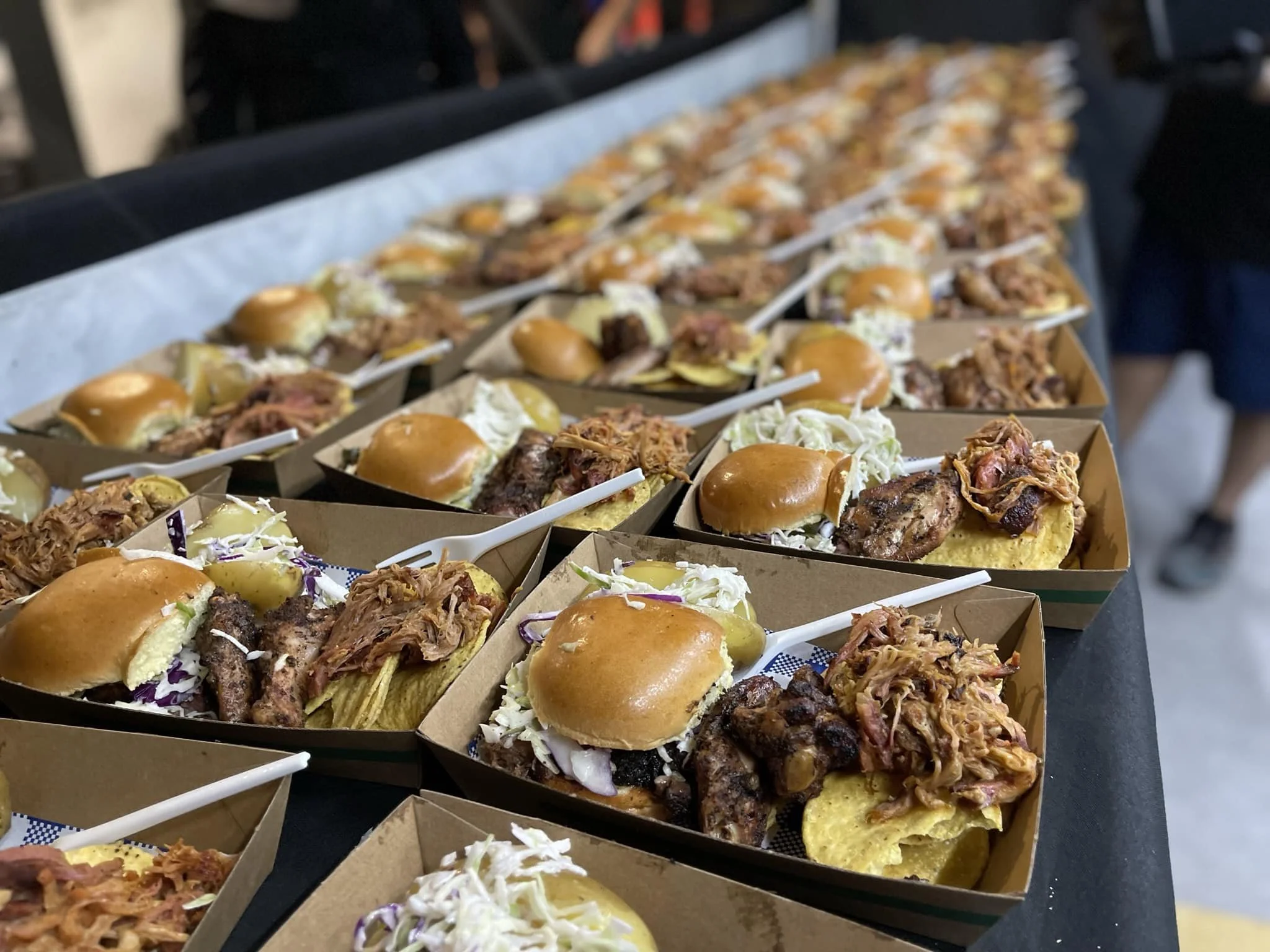 Tray of mini sandwich plates with pulled pork, cole slaw, pickles, and buns arranged on a table.