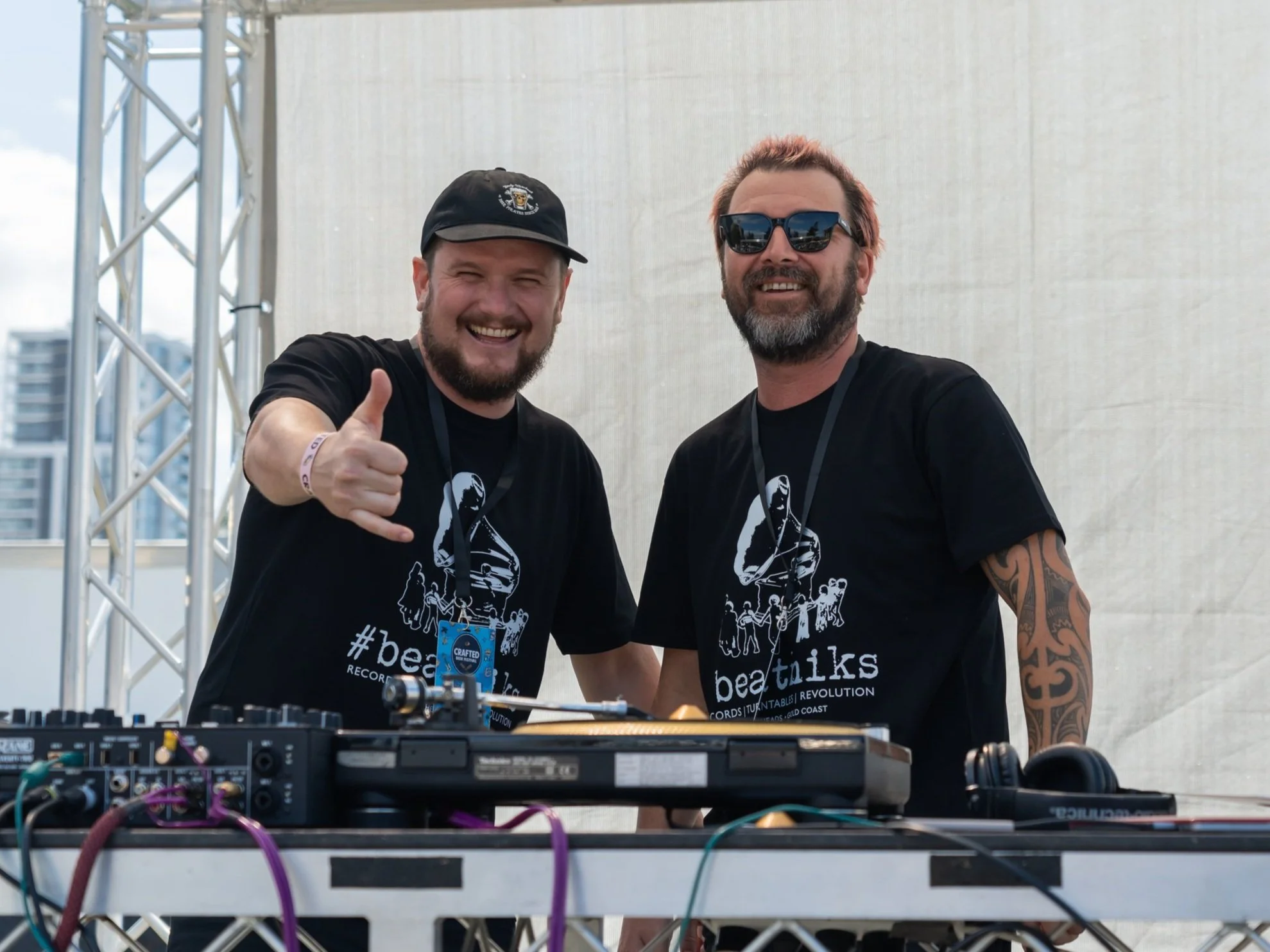 Two smiling men wearing black shirts with 'beatniks' logo, standing behind DJ equipment on a stage outdoors. One man is giving a thumbs-up and the other is wearing sunglasses. The background shows a beige wall and parts of a metal stage structure.