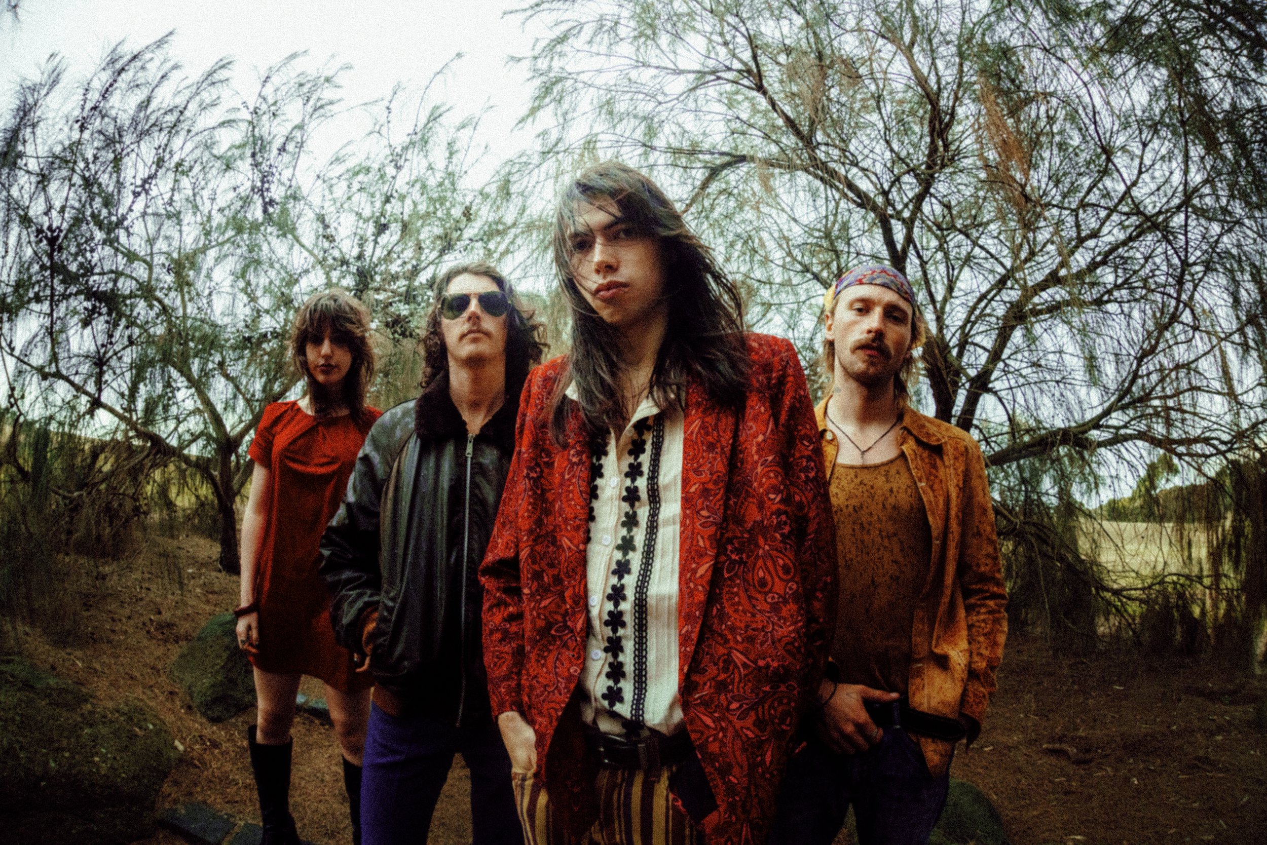 A group of five young adults standing outdoors in front of leafless trees, dressed in vintage and bohemian clothing, with serious expressions.