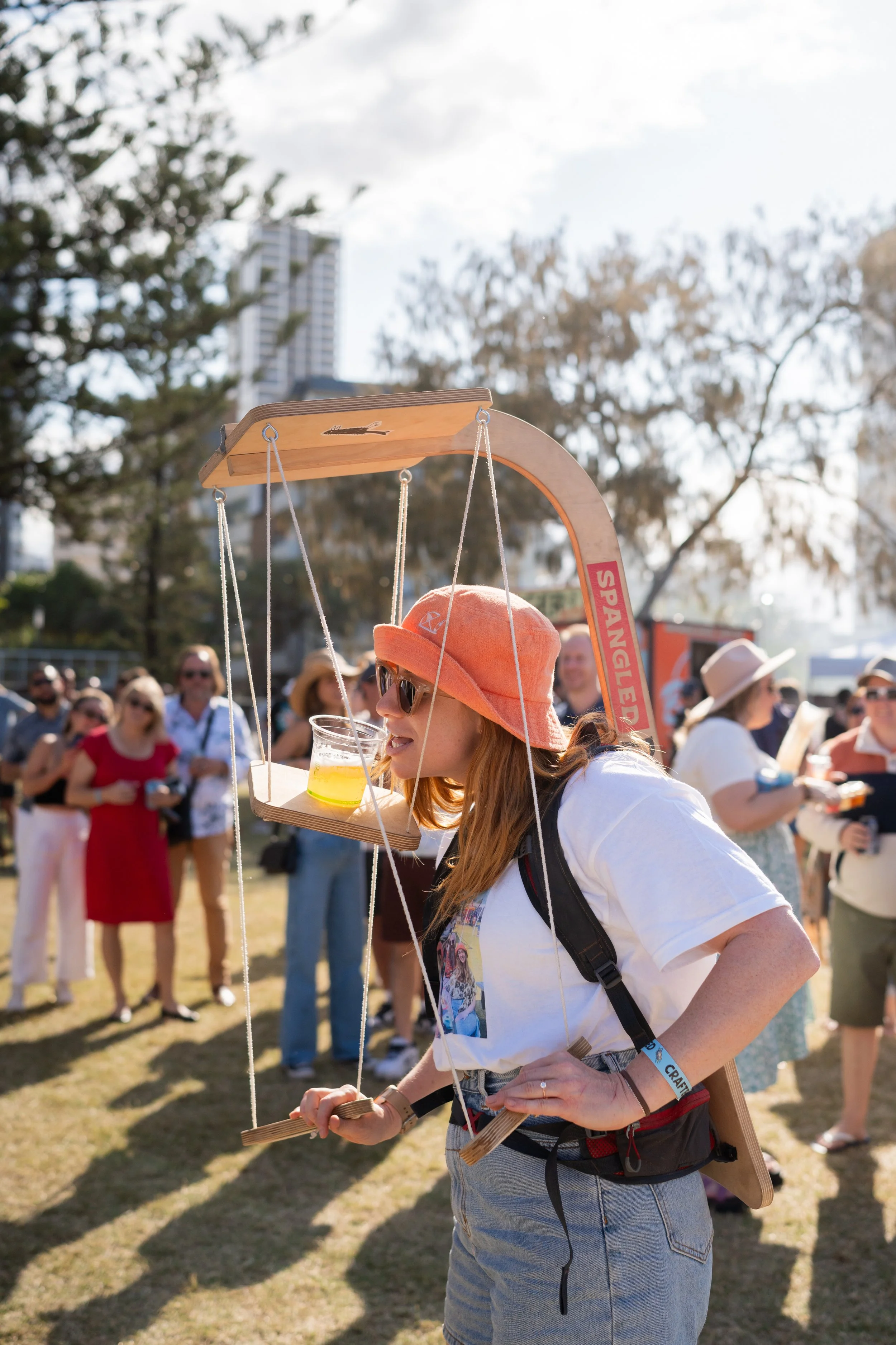 A woman with orange hat and sunglasses carrying a wooden balance scale with a plastic cup filled with yellow liquid, at an outdoor event with people in the background.