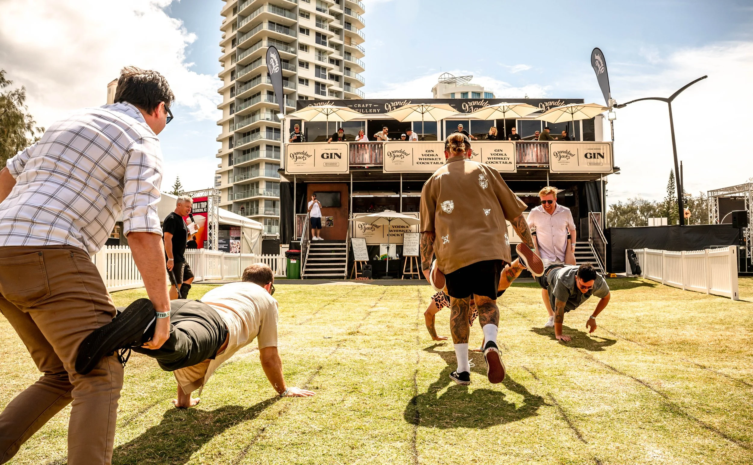 People doing push-ups on grass outdoors during daytime at event with a two-story bar or pub structure behind them, with tall buildings in the background.