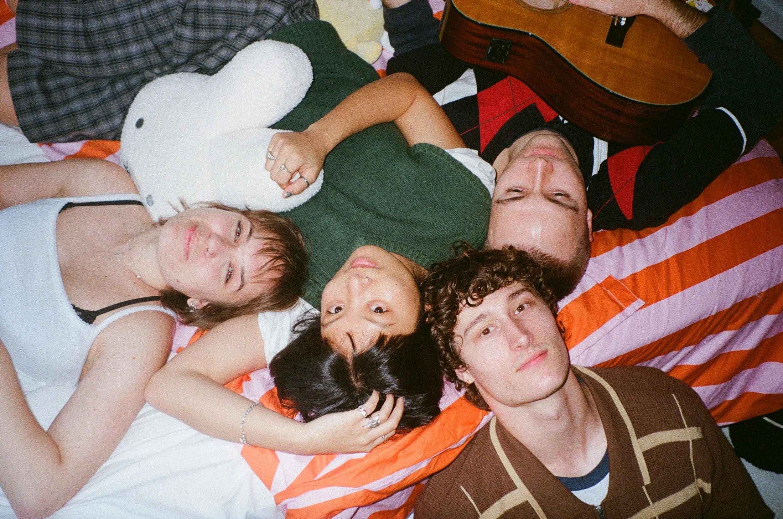 Group of four friends lying on a bed, their heads close together, one holding a guitar, with a stuffed animal and colorful striped bedding.