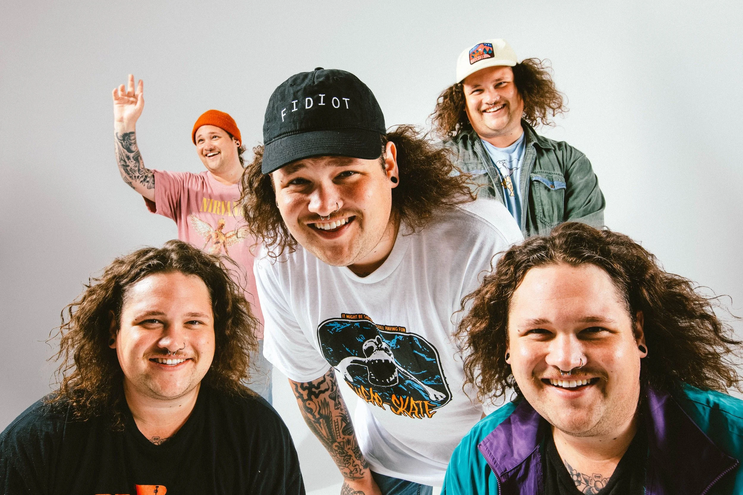 Group of six happy men with various tattoos and piercings, smiling and posing together in front of a plain white background. They are wearing casual clothing, including t-shirts, jackets, and hats.