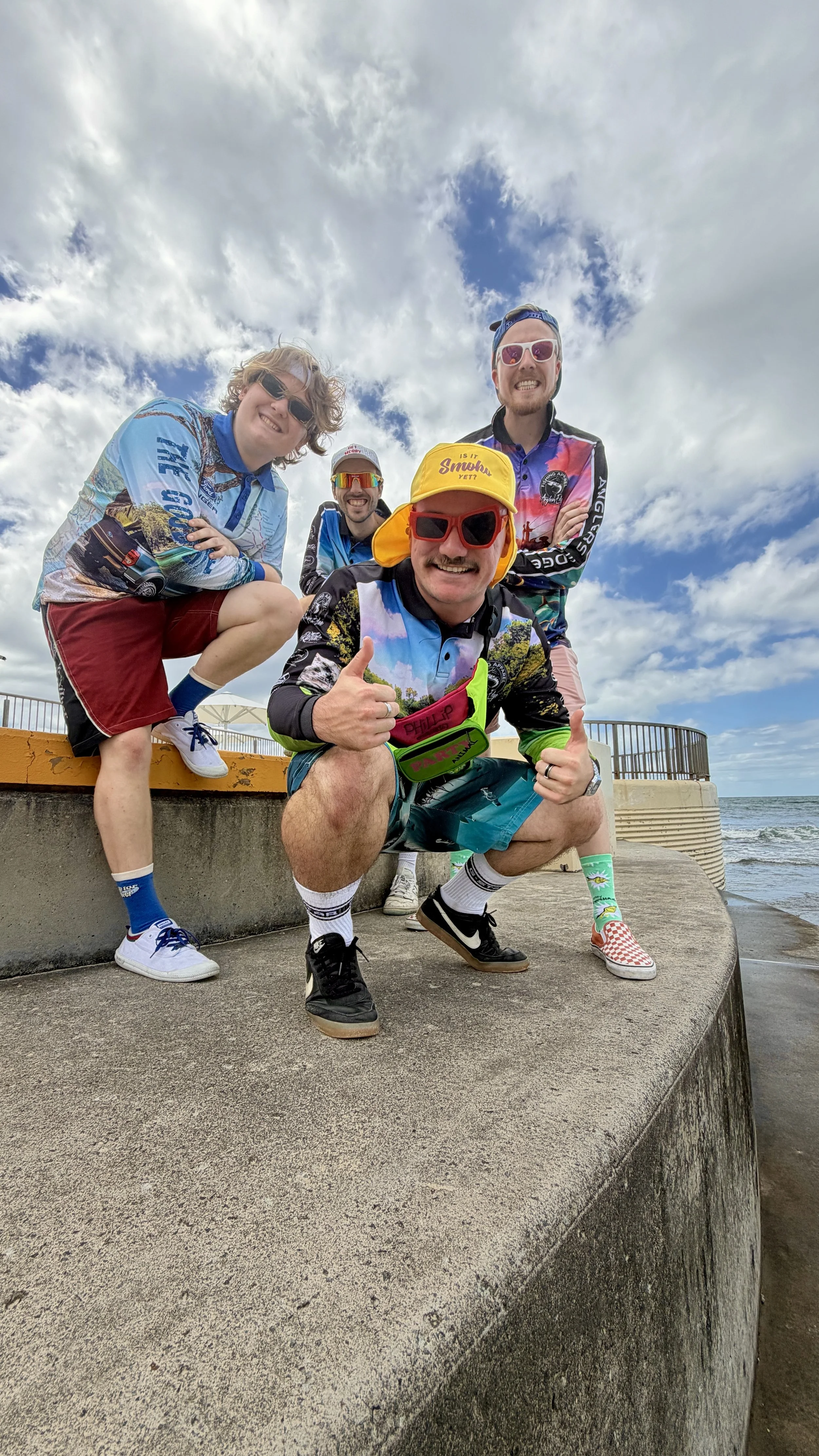Four men dressed in colorful casual and beachwear clothing squatting on a concrete drain cover near the beach, smiling and posing with thumbs up under a partly cloudy sky.