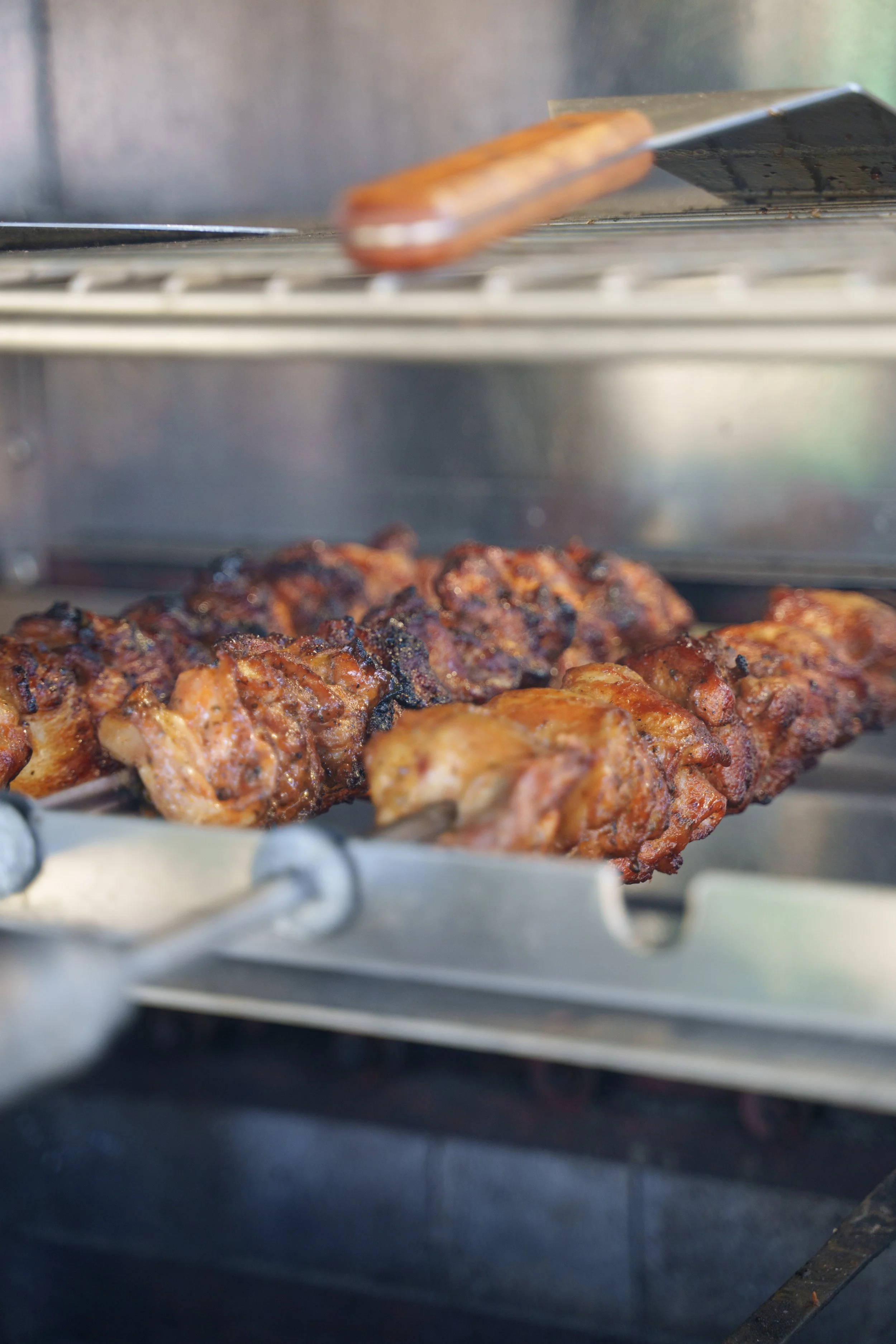 Close-up of skewered grilled meat, possibly chicken, on a rotisserie in a stainless steel grill or oven with a metal rack above.