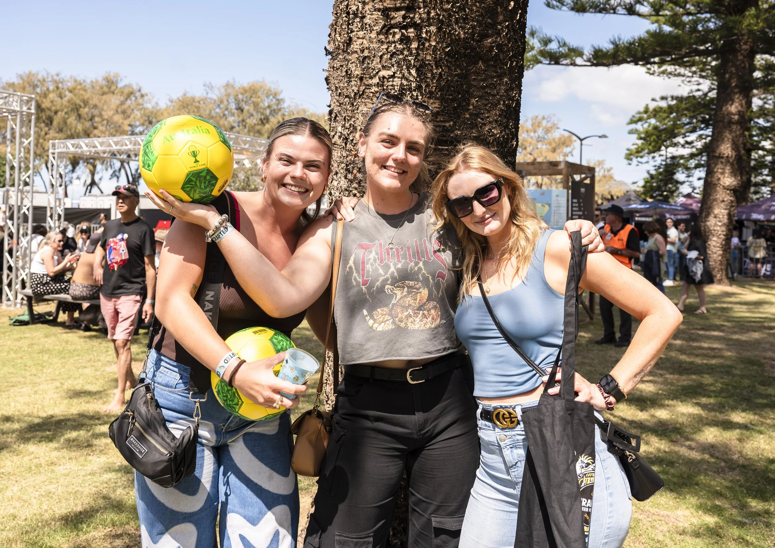Three women smiling and posing together outdoors at a festival or event during the daytime, with one woman holding a yellow and green soccer ball.