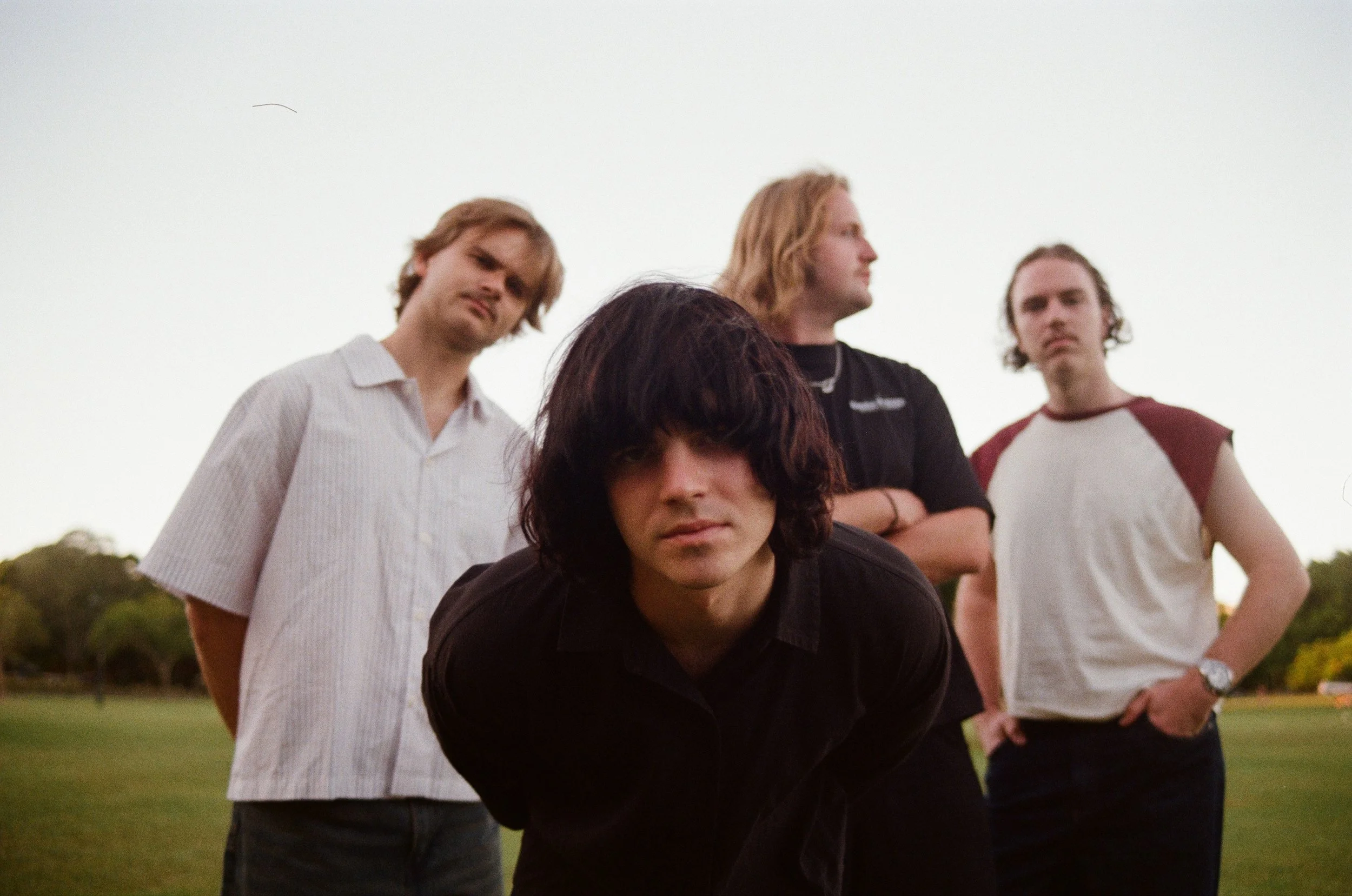 A group of five young men standing outdoors on a grassy field during sunset, with three of them visible, two in the back and one in front. They have various hair colors and styles, with serious expressions, and are dressed casually.