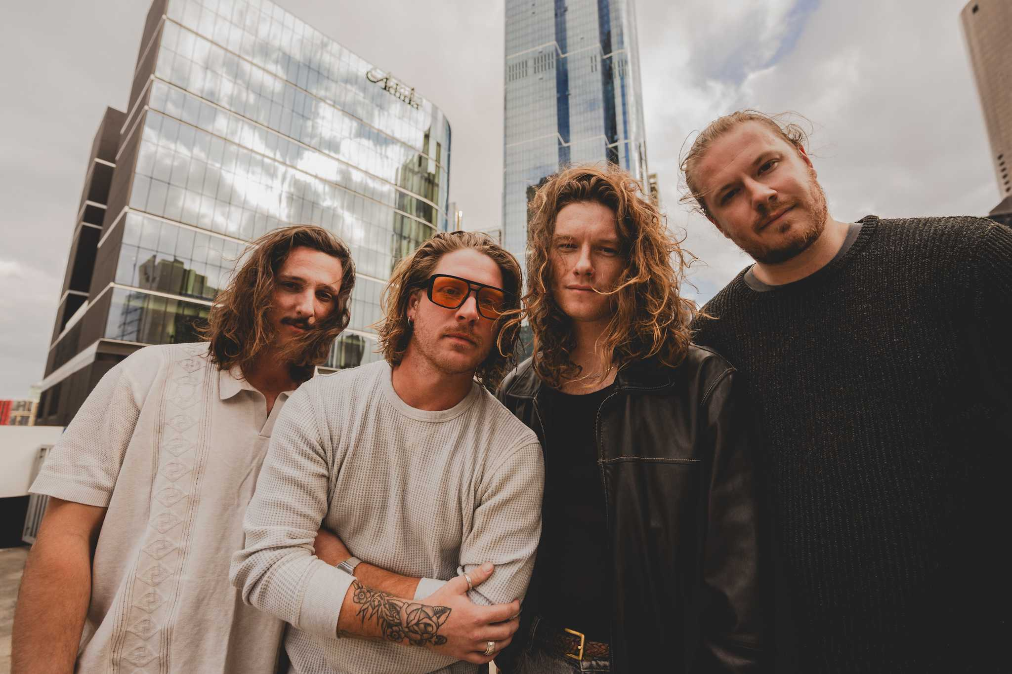 A group of four young adults, three men and one woman, standing close together outdoors in an urban setting with modern glass skyscrapers in the background. The woman has curly hair, wears sunglasses, and is dressed in a black jacket. The men have long hair, with one man having a tattoo on his forearm.