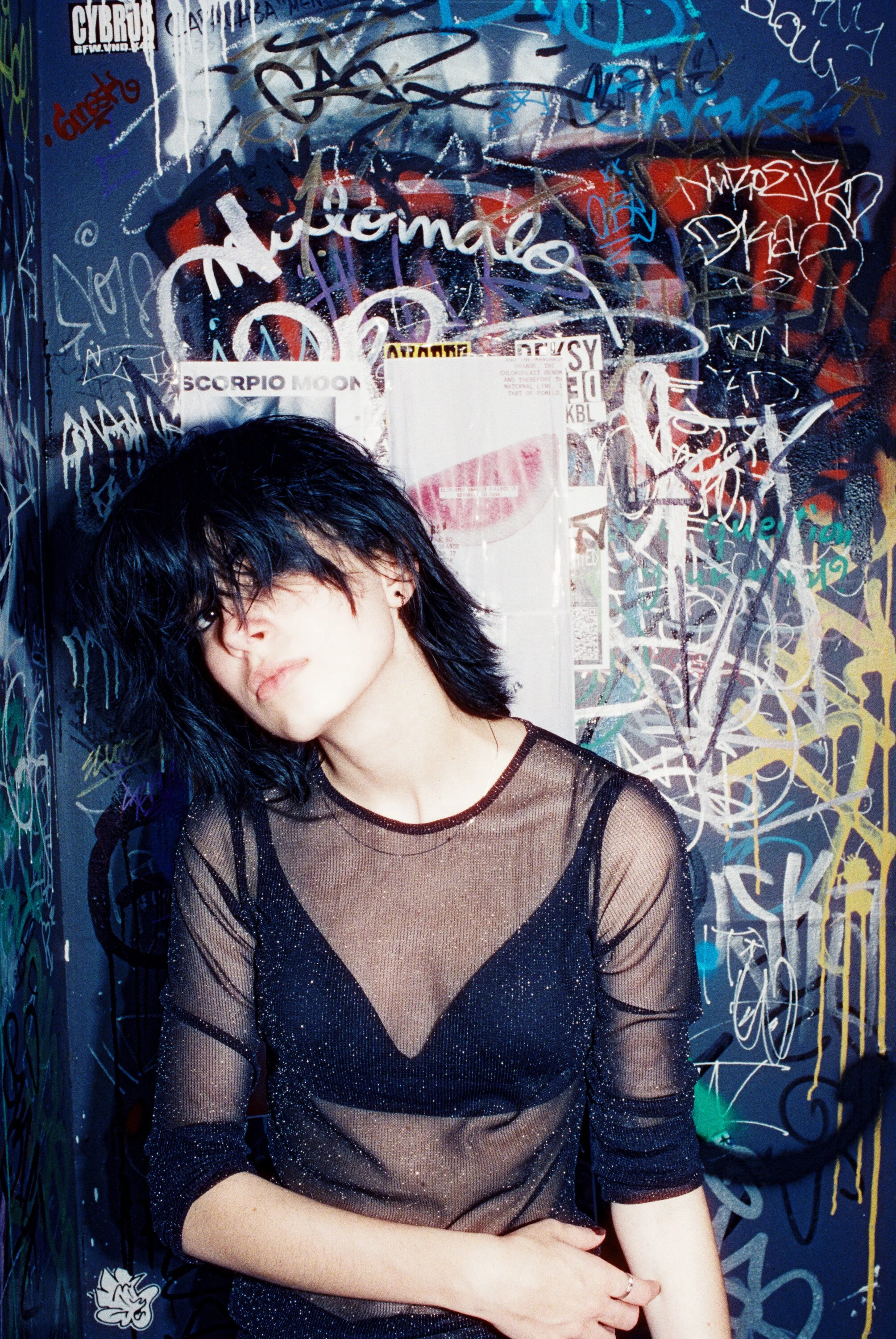 A woman with black, shaggy hair covering part of her face, wearing a sheer black glittery top over a black bra, standing in front of a graffiti-covered wall.